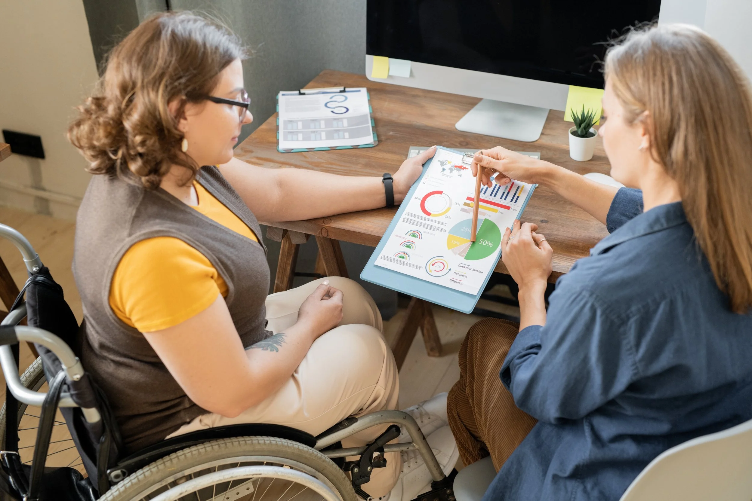 Two women discussing a document with graphs and charts, one in a wheelchair, at a wooden desk with a computer monitor and plant.