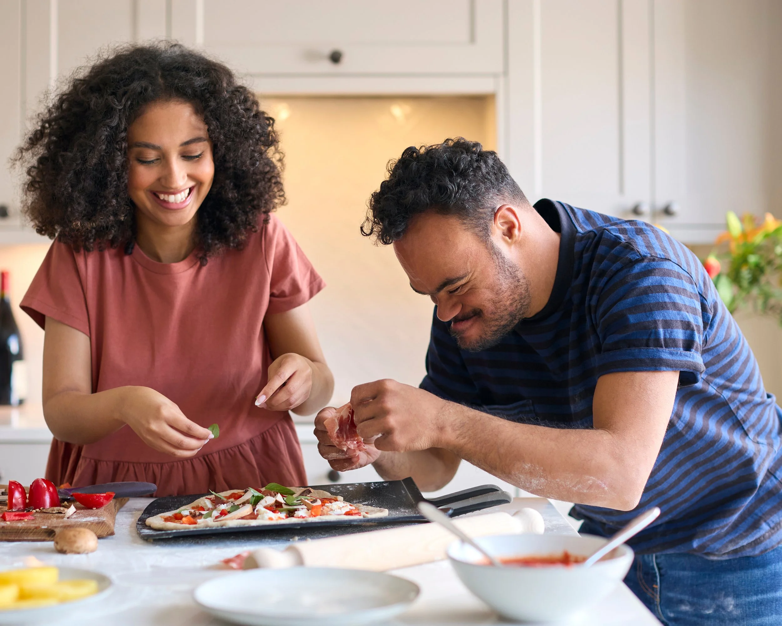 A smiling couple making pizza in a kitchen, adding toppings to dough. A bowl of sauce and sliced ingredients are on the counter.