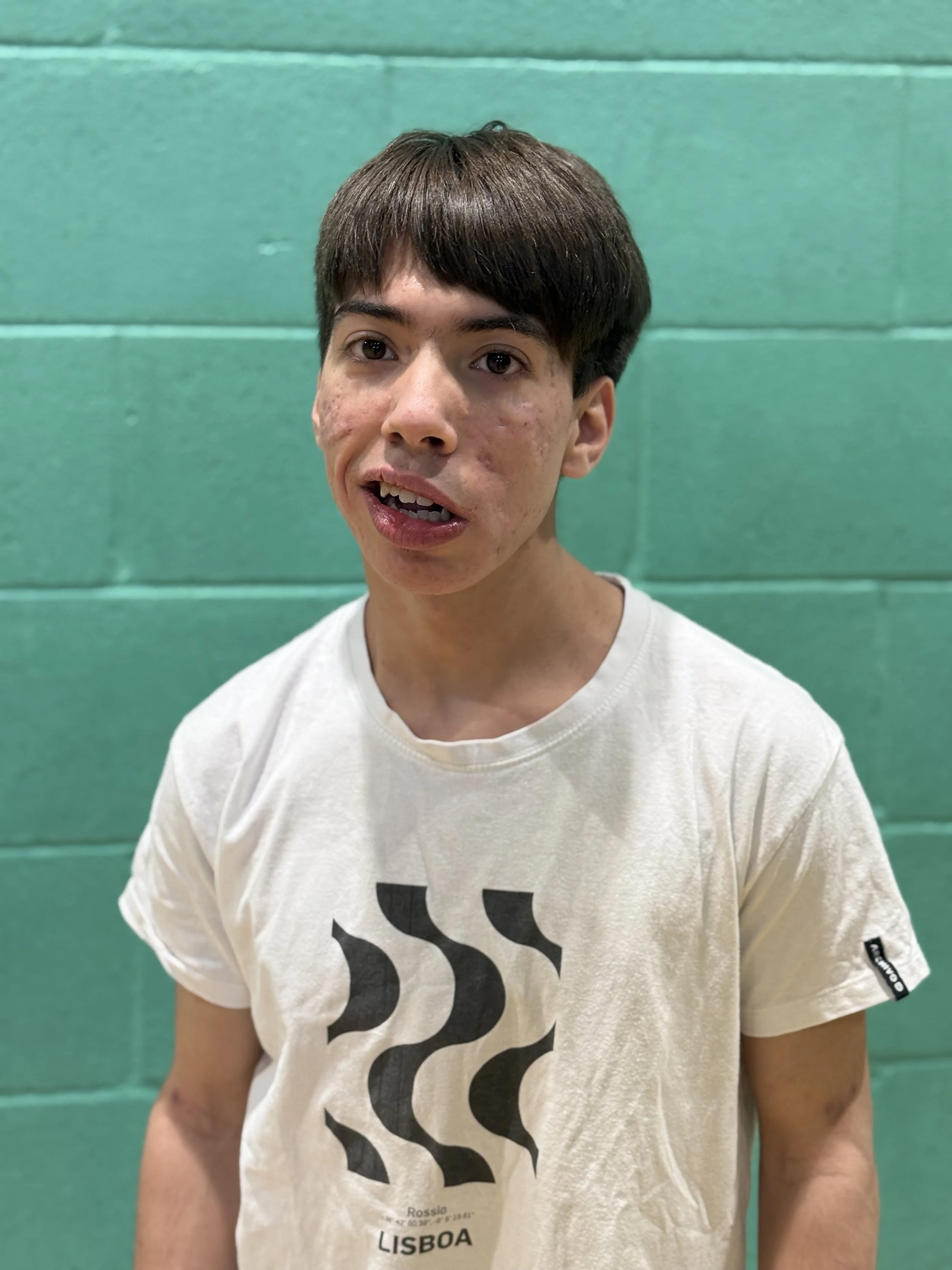 Young volunteer in a white t-shirt standing in front of a sports hall wall, looking directly at the camera with a neutral expression.