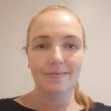 Portrait of Meagan Rees. She is a woman with blonde hair tied back, wearing a light necklace and a neutral top, standing against a plain light background.