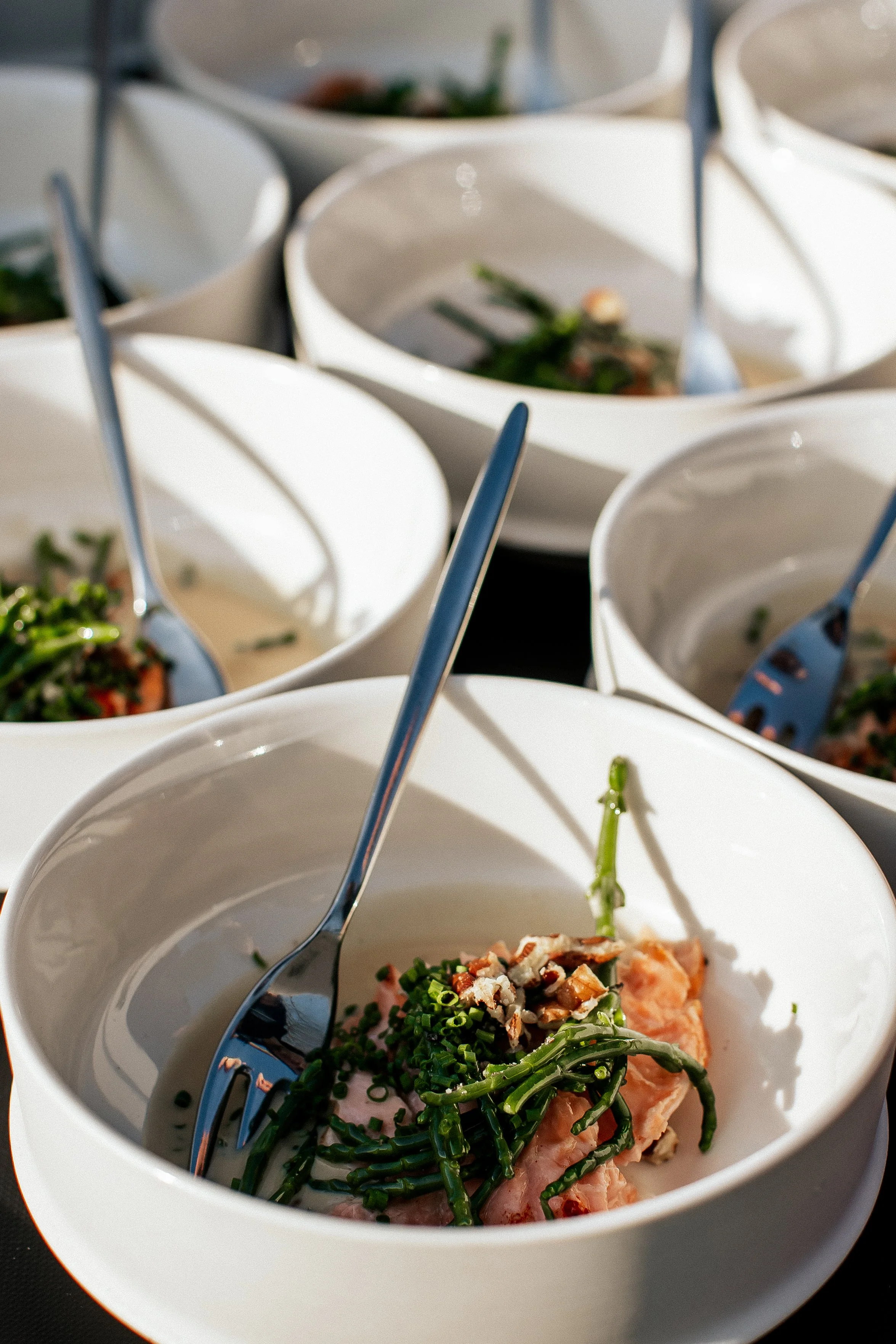 White bowls containing salmon, green vegetables, nuts, and forks, arranged on a table.
