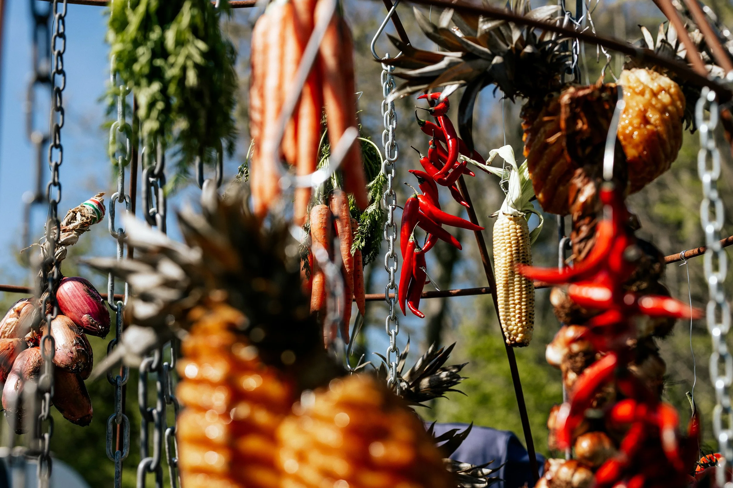 Hanging vegetables and fruits including carrots, red chili peppers, onions, corn, and pineapples.