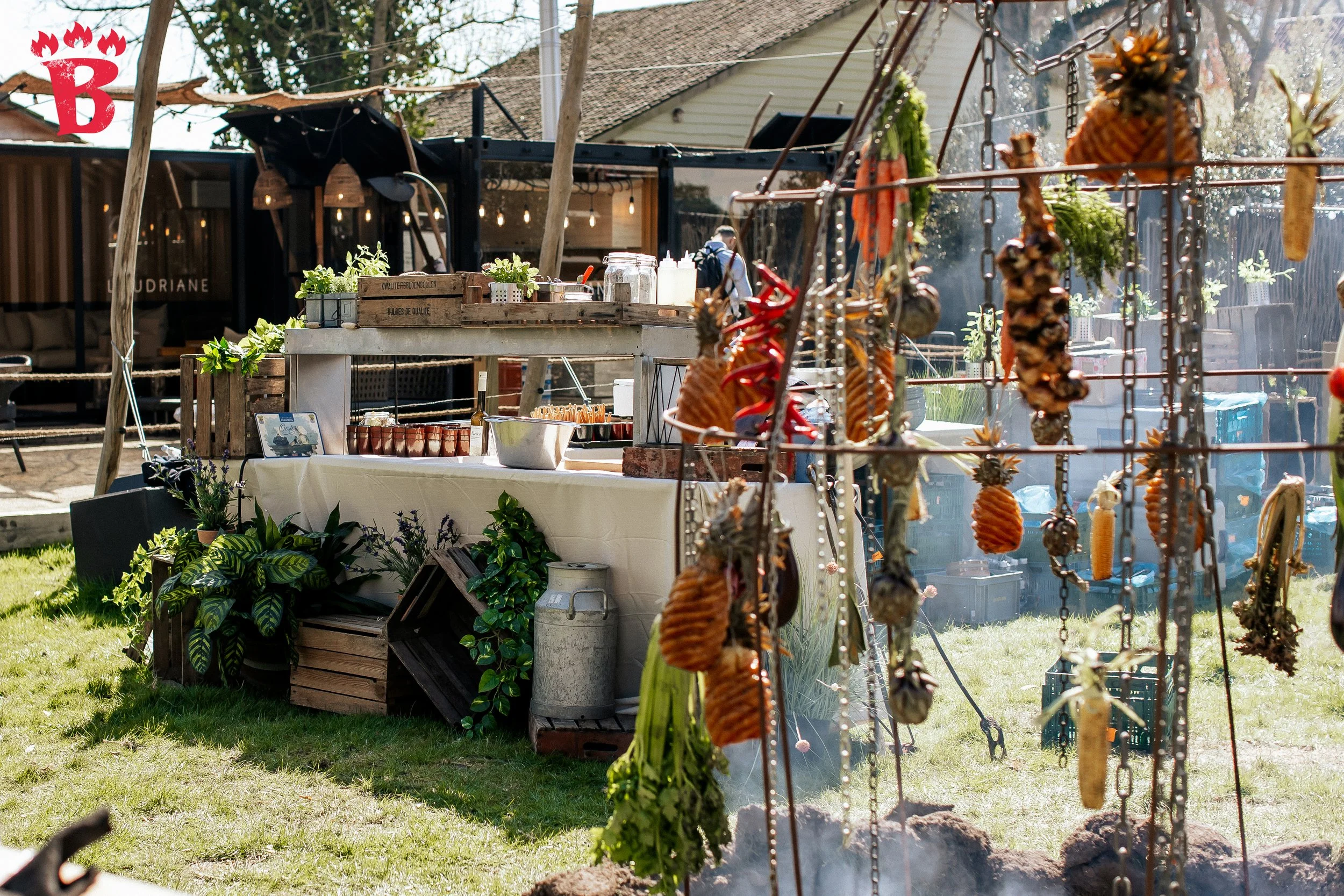 Outdoor cooking setup with pineapples, peppers, and vegetables hanging over a grill; nearby table with plants and kitchen items.