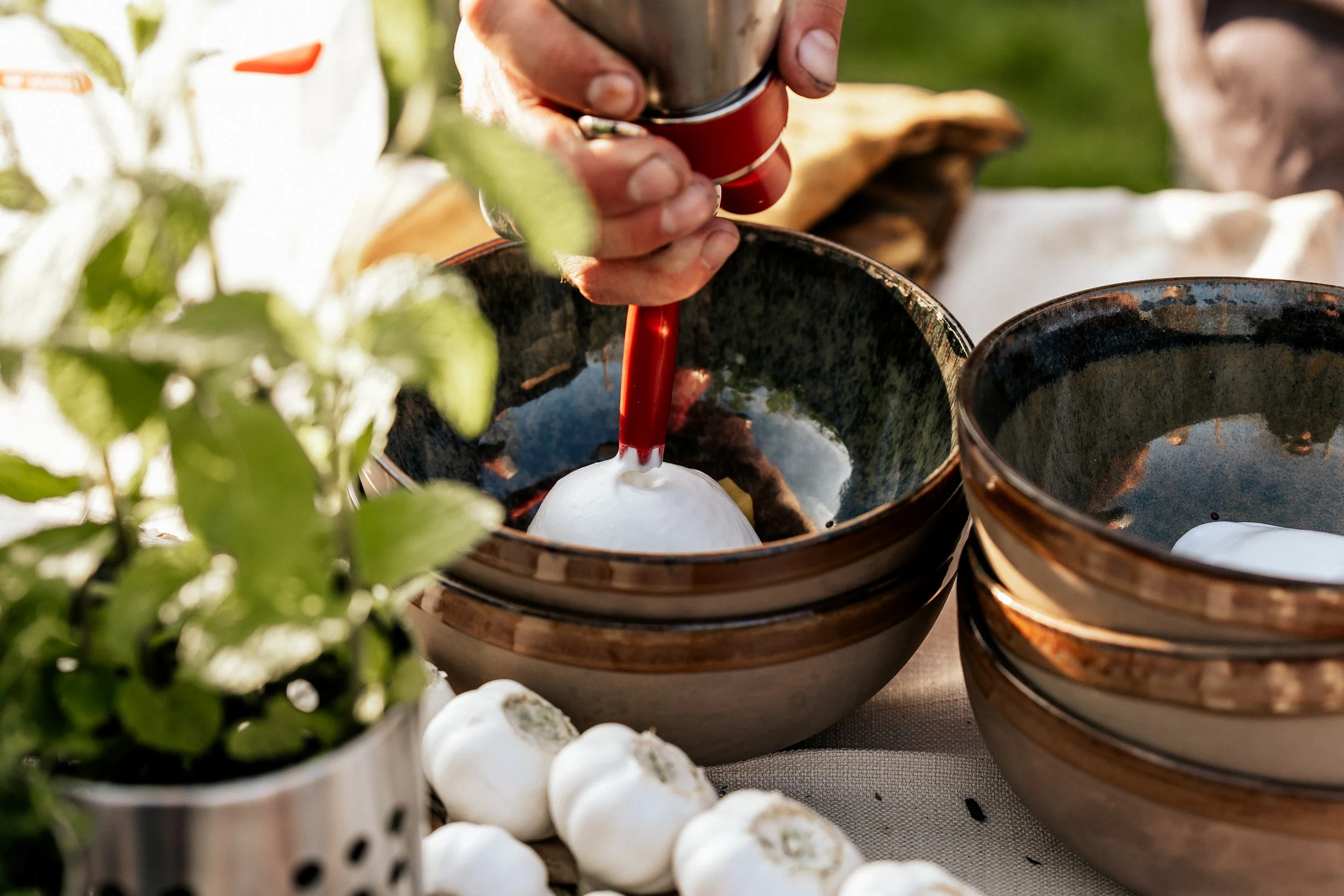 Hand piping foam into ceramic bowl outdoors, with herbs and garlic in the background.