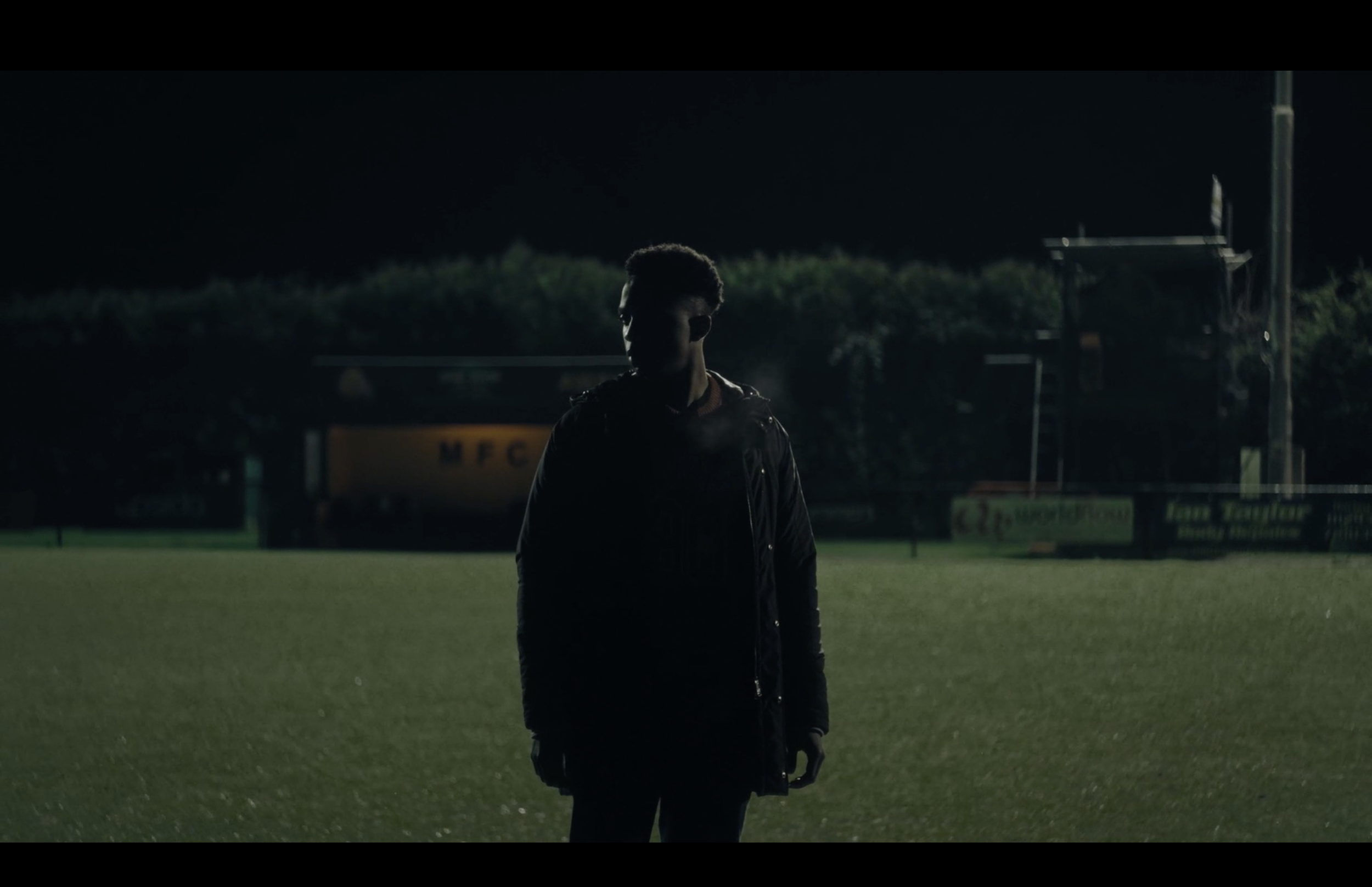 A person standing alone, in the dark, on a football pitch.