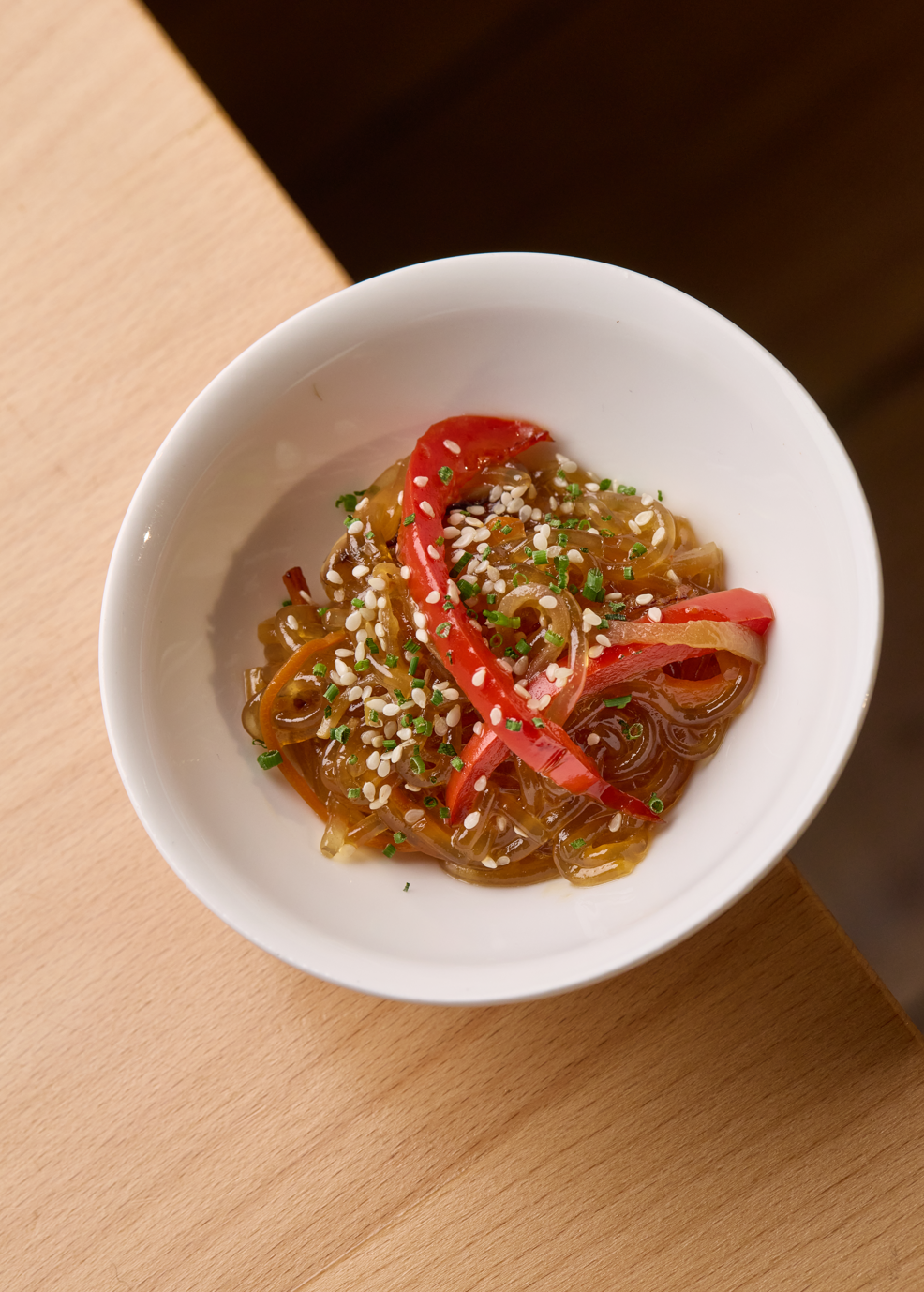 Un bol blanc contenant des nouilles de style asiatique, garnies de poivrons rouges, de graines de sésame et d'herbes vertes, placé sur une surface en bois.