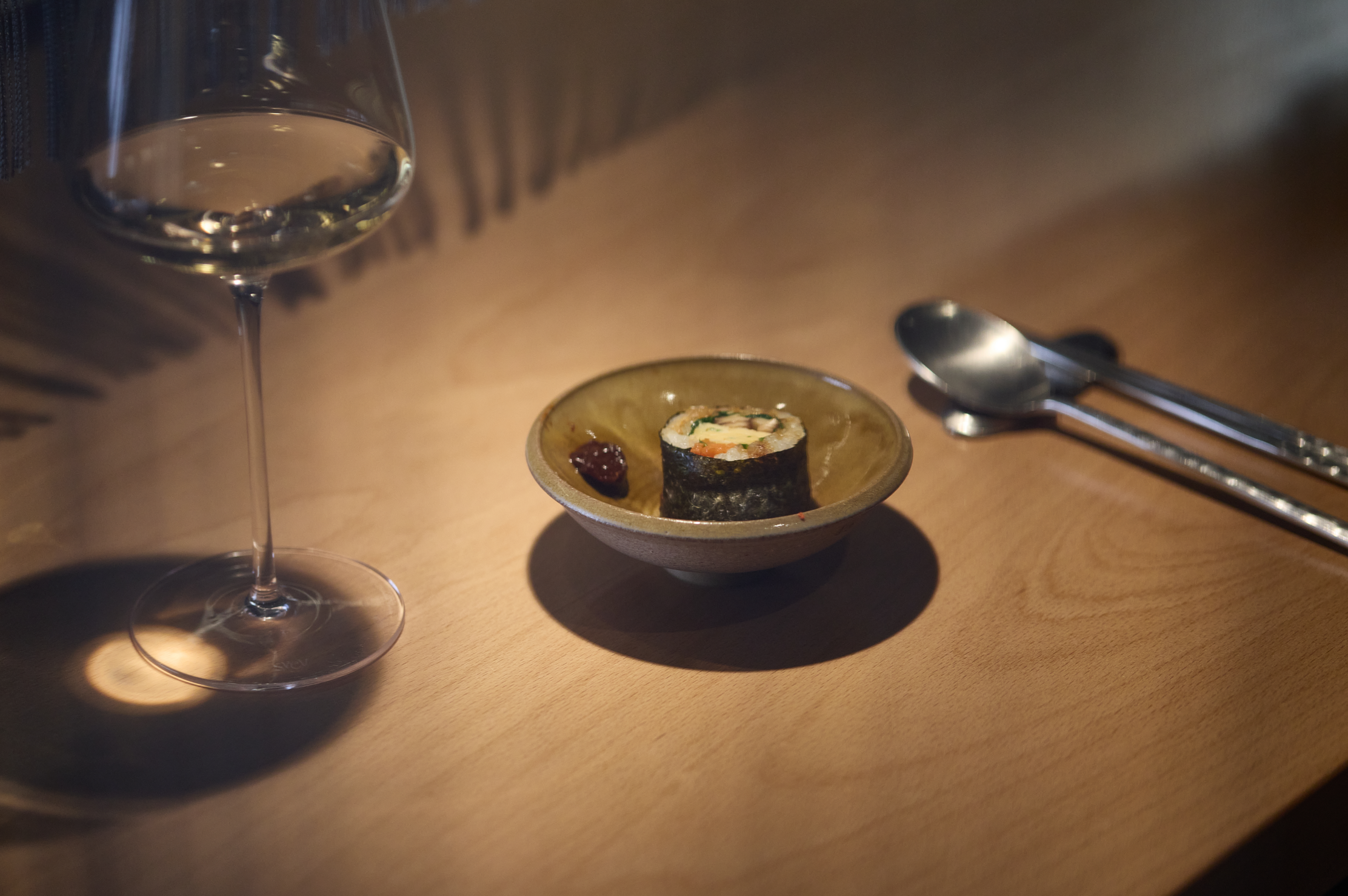 Un verre d'eau, un petit plat avec un rouleau de sushi et une petite sauce, une cuillère et une paire de baguettes posés sur une table en bois.