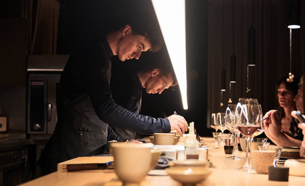 Deux chefs en train de préparer des plats dans une cuisine professionnelle, avec un groupe de personnes regardant et prenant des notes ou des photos.