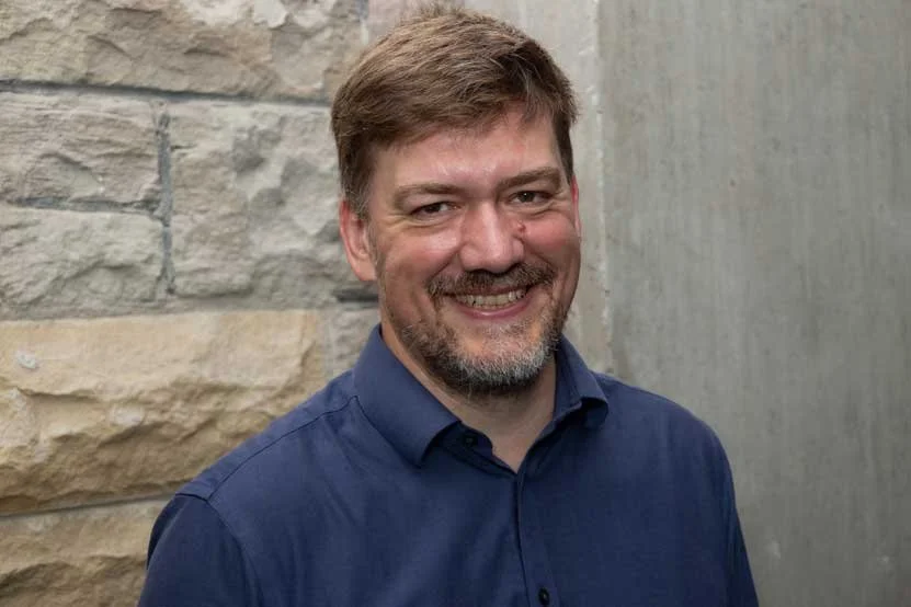 Iain Tinsdale- A smiling man with brown hair and a beard wearing a navy blue collared shirt, standing against a textured stone and concrete wall.