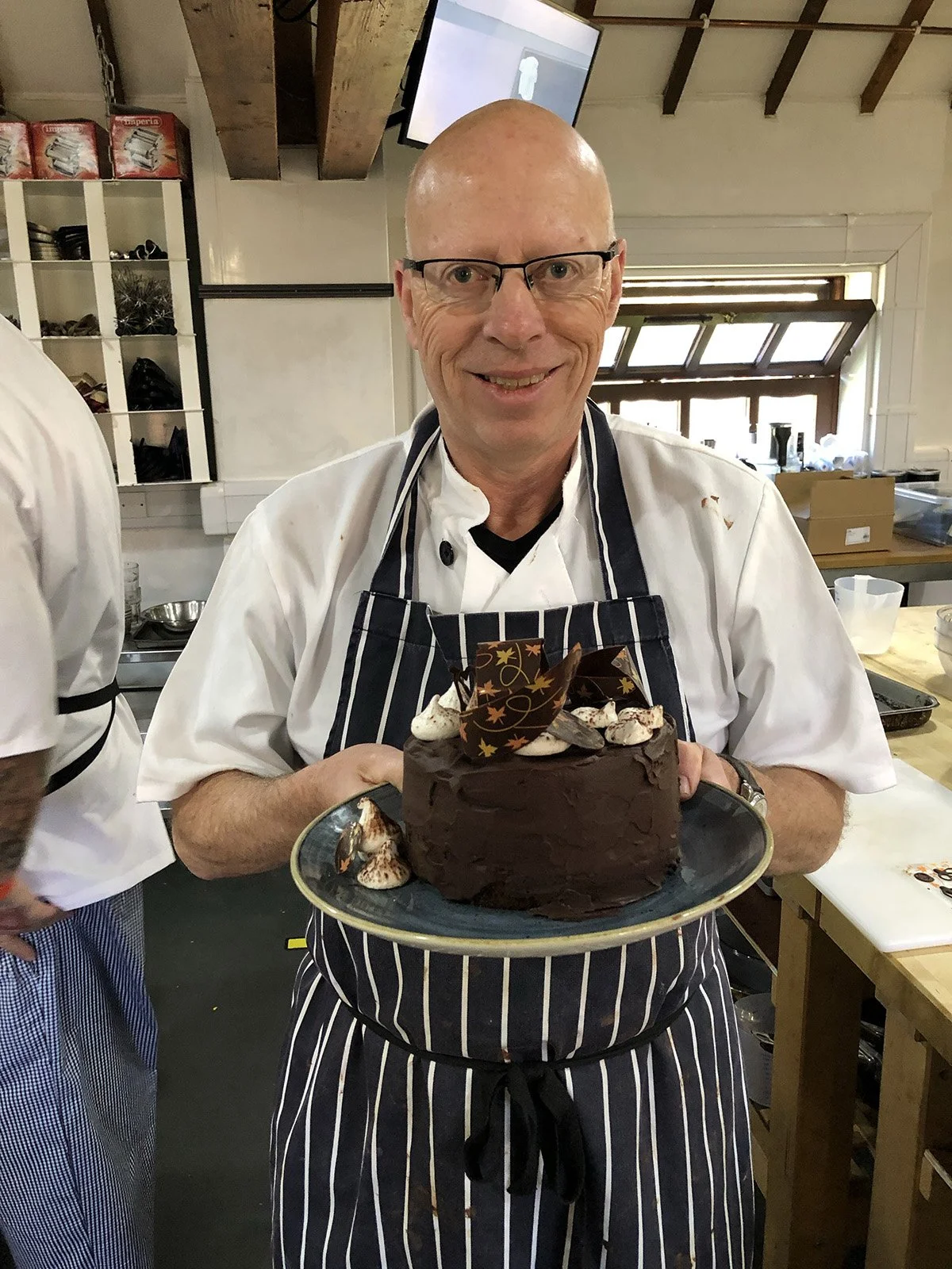 Chef Colm, holding a chocolate cake decorated with chocolate pieces and whipped cream in a kitchen.