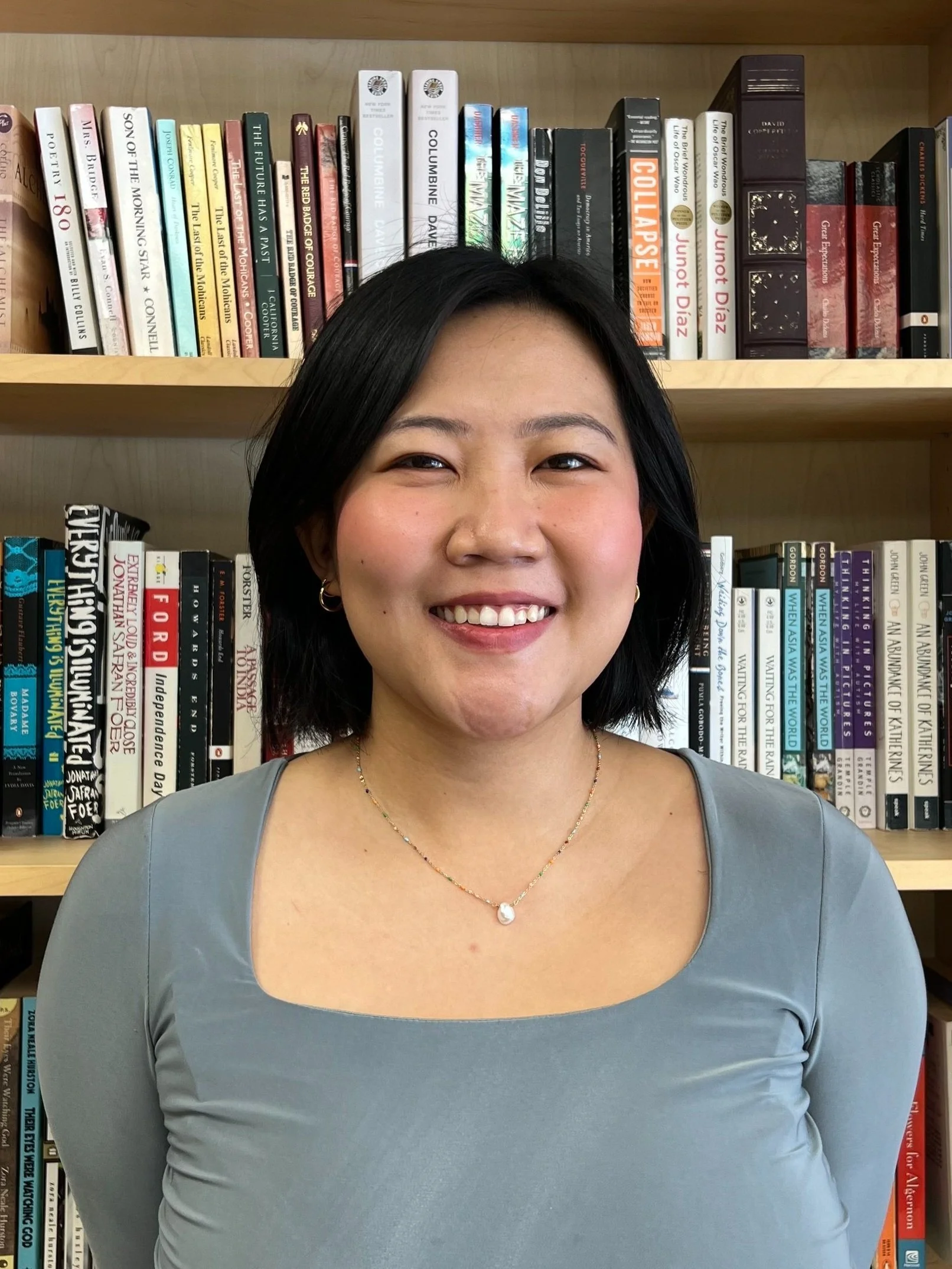 A woman with short black hair smiling, wearing a gray top and a necklace, standing in front of a bookshelf filled with various books.