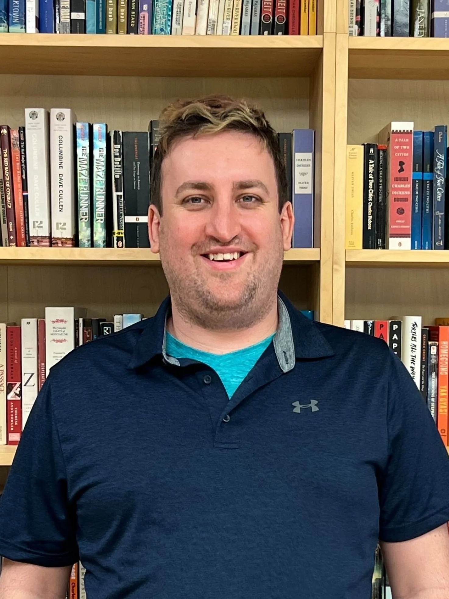 Smiling man in navy blue polo shirt standing in front of a wooden bookshelf filled with various books.