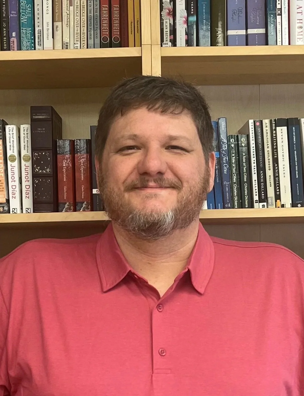 A man with short brown hair, a beard, and a mustache, wearing a pink collared shirt, standing in front of a bookshelf filled with books.