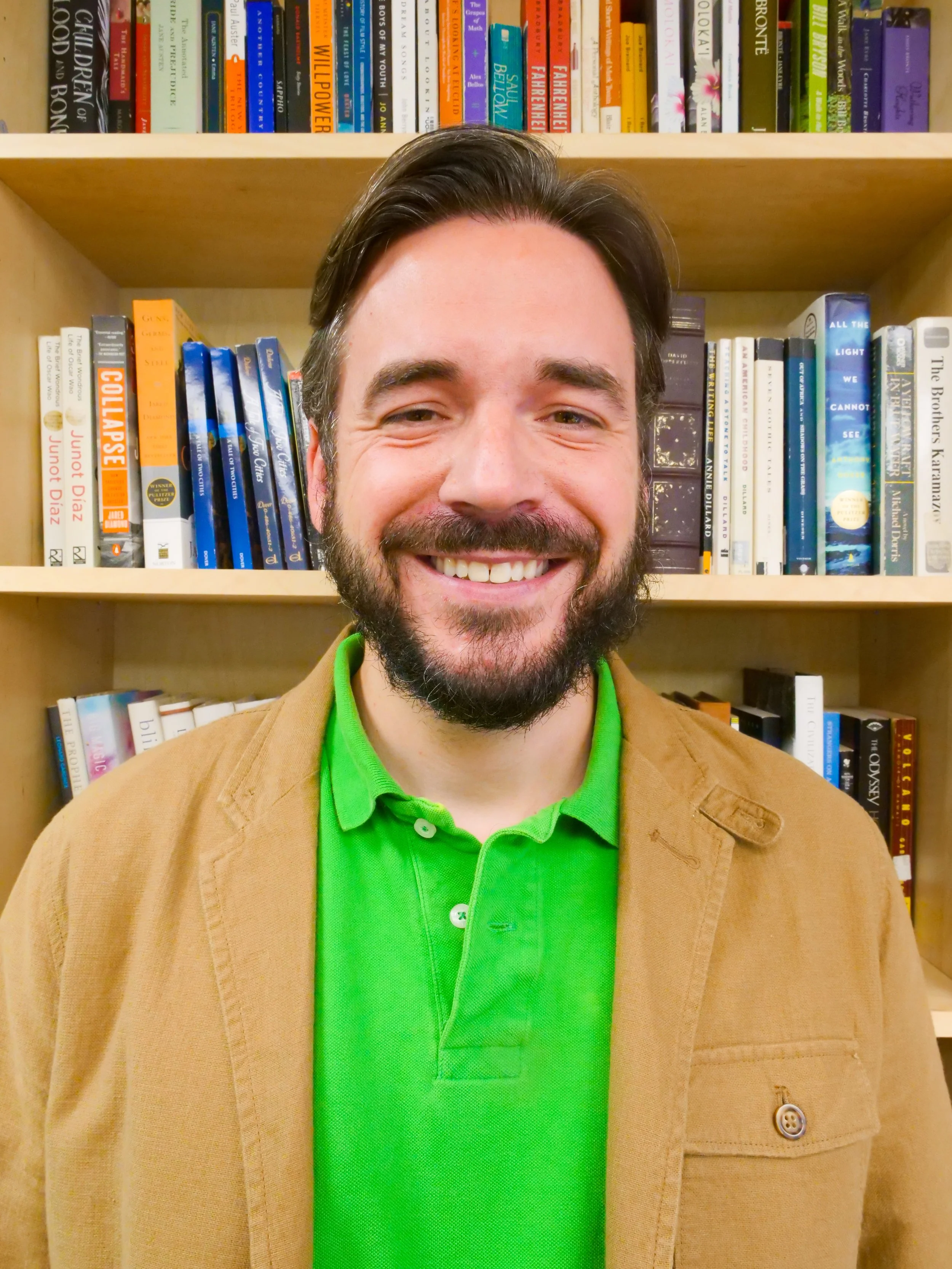 Portrait of a smiling man with dark hair and a beard, wearing a bright green polo shirt and a tan jacket, standing in front of a bookshelf filled with colorful books.