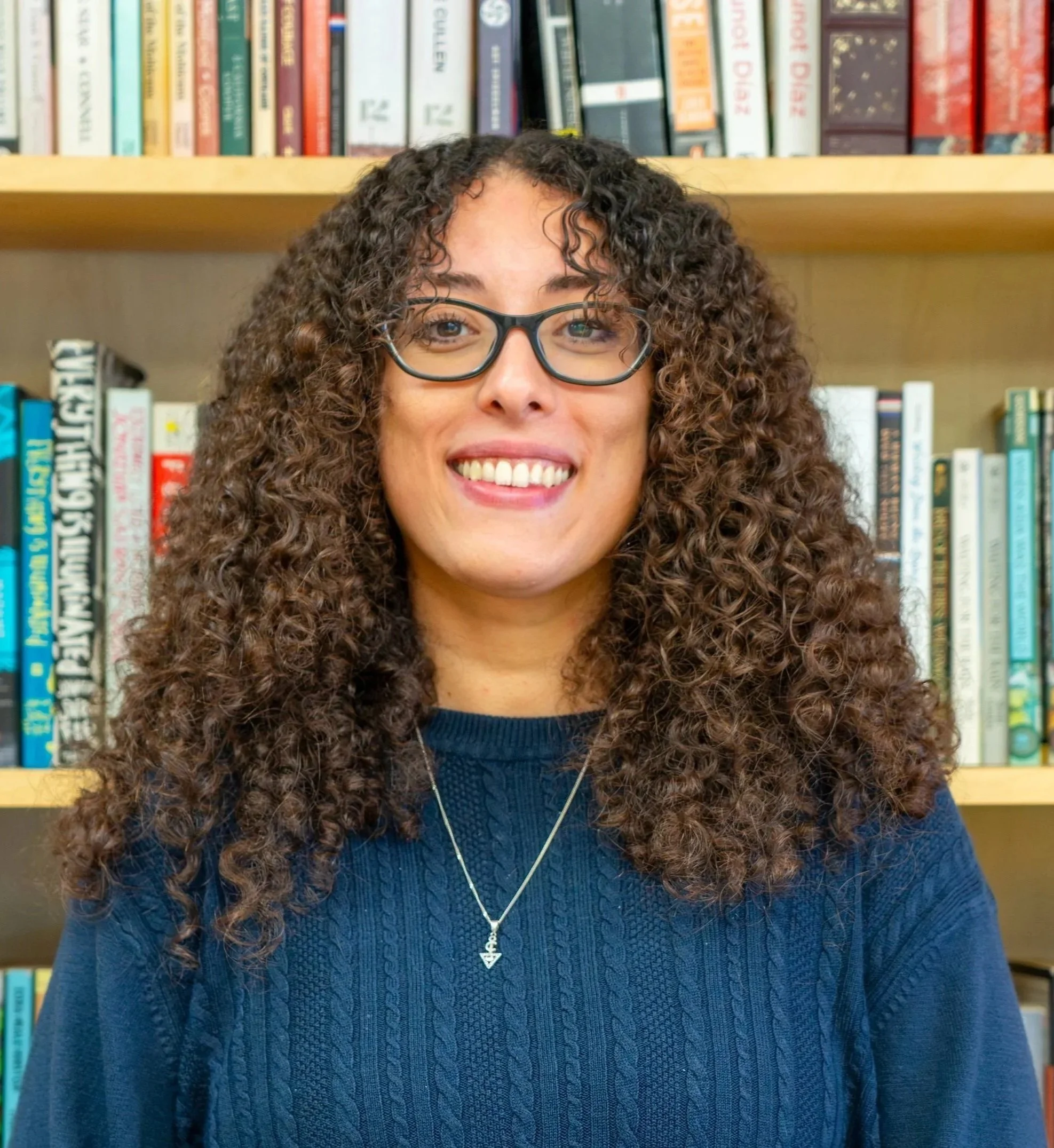 A woman with curly hair and glasses smiling in front of a bookshelf filled with books.