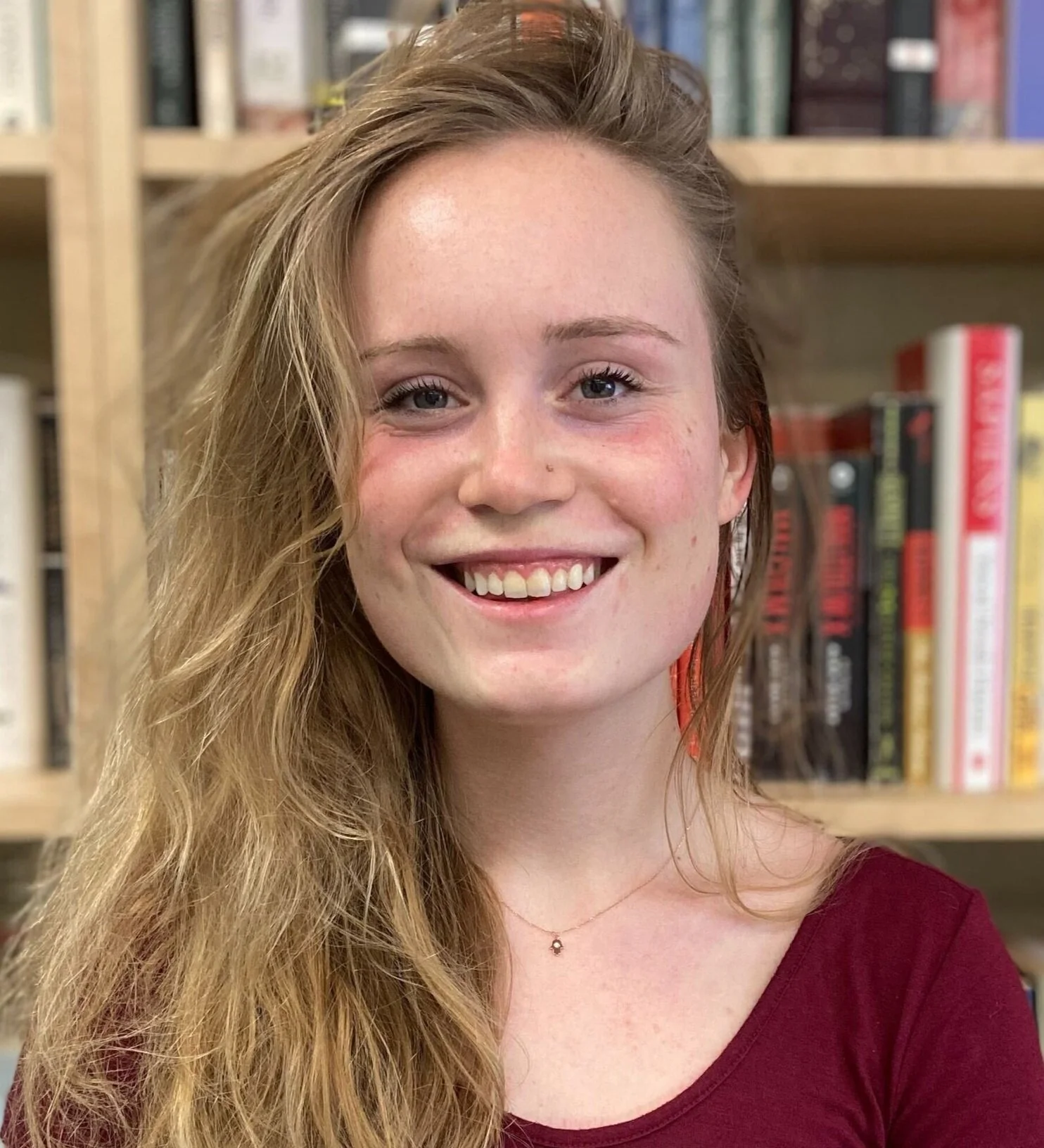 A smiling young woman with long, wavy blonde hair and a nose piercing, wearing a maroon shirt and a delicate necklace, standing in front of a bookshelf filled with books.