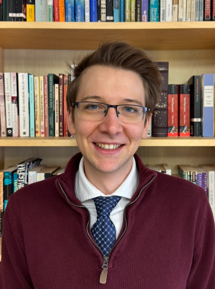 A young man with glasses, smiling, wearing a maroon zip-up sweater over a collared shirt and tie, with bookshelves in the background.