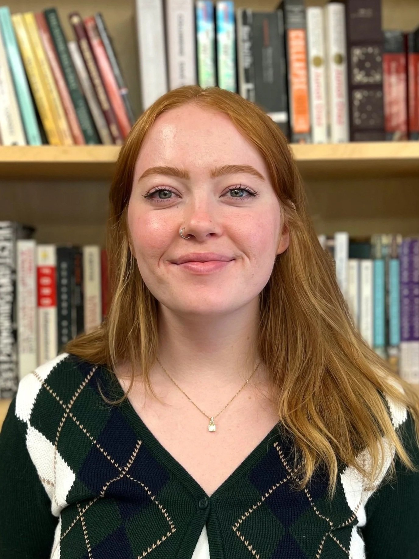 A woman with red hair, blue eyes, and a nose piercing, smiling, standing in front of a bookshelf filled with various books.