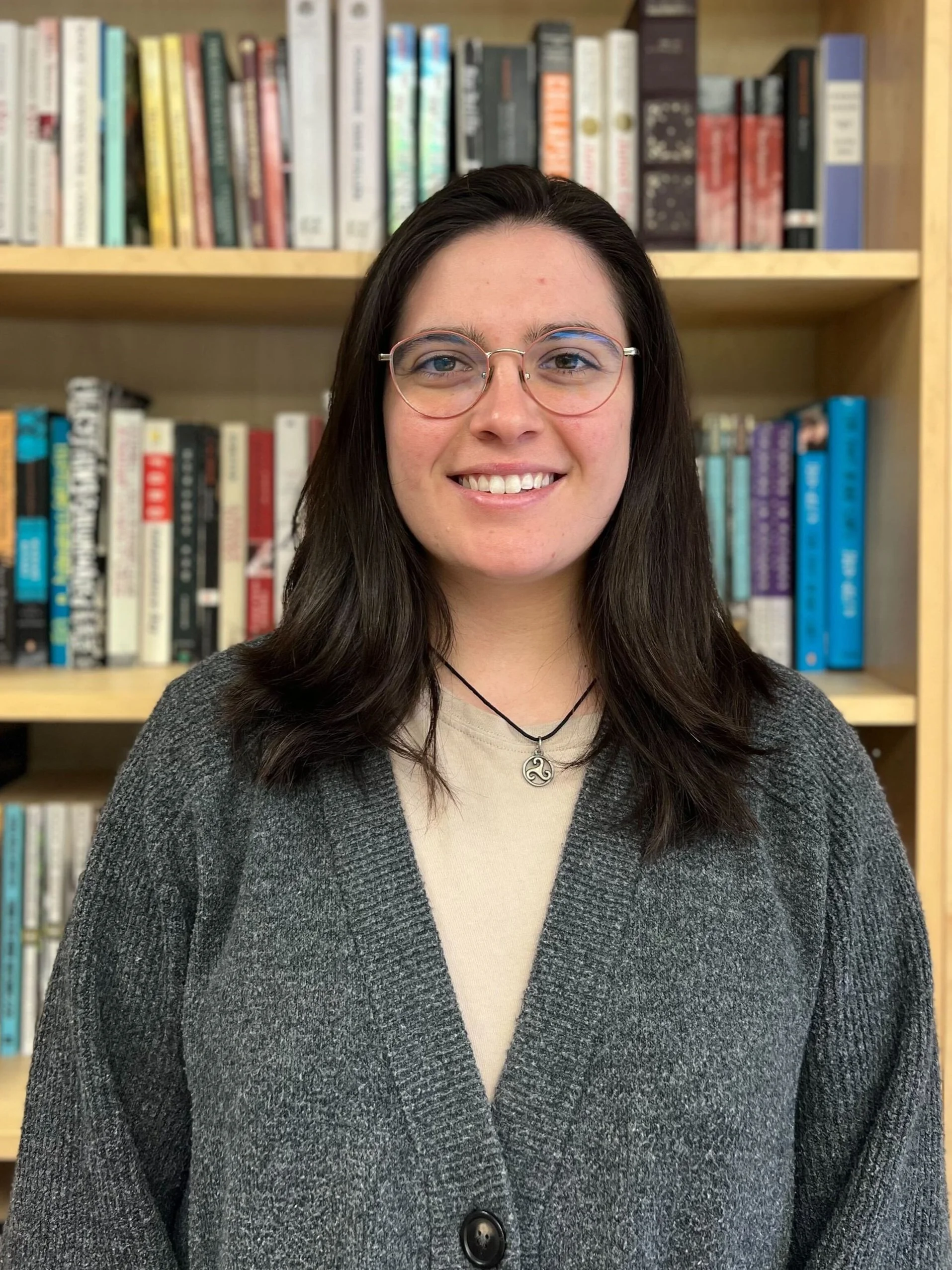 A young woman with glasses and dark brown hair smiling in front of a bookshelf filled with colorful books.