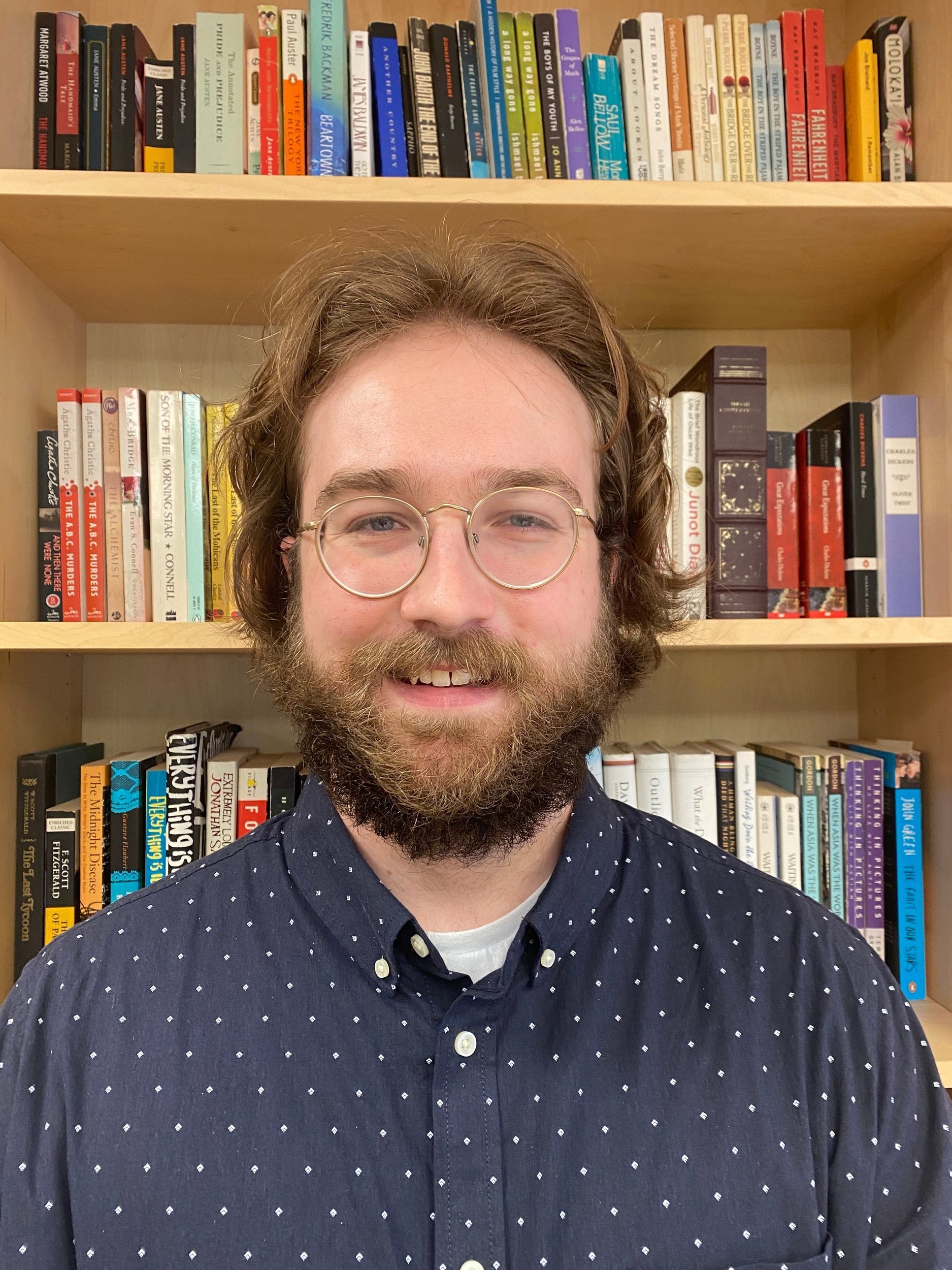 A bearded man with glasses smiling in front of bookshelves filled with various books.