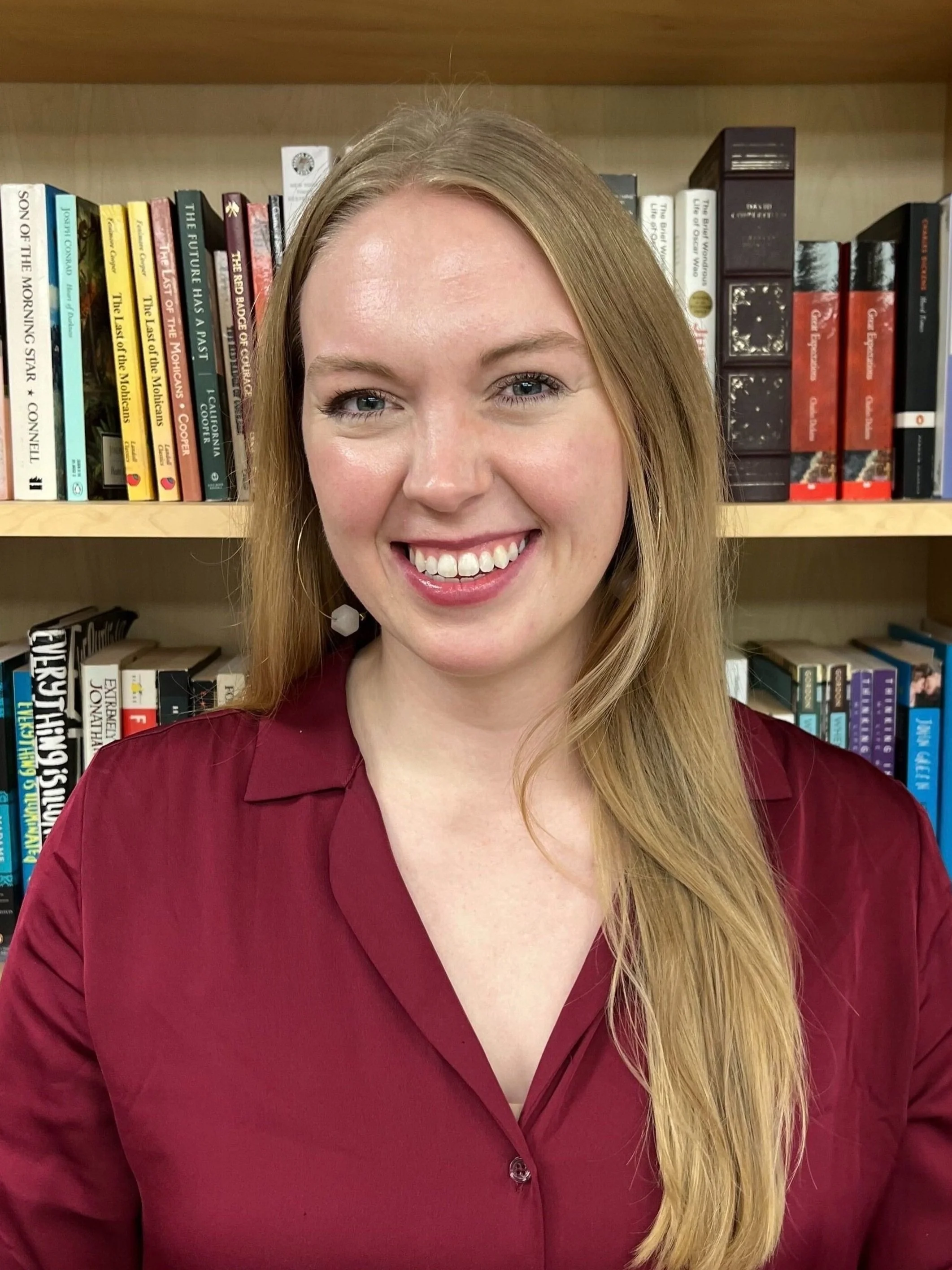 A woman with long blonde hair smiling in front of bookshelf filled with books.