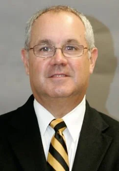 Man in suit with glasses and a striped tie, smiling against a neutral backdrop.