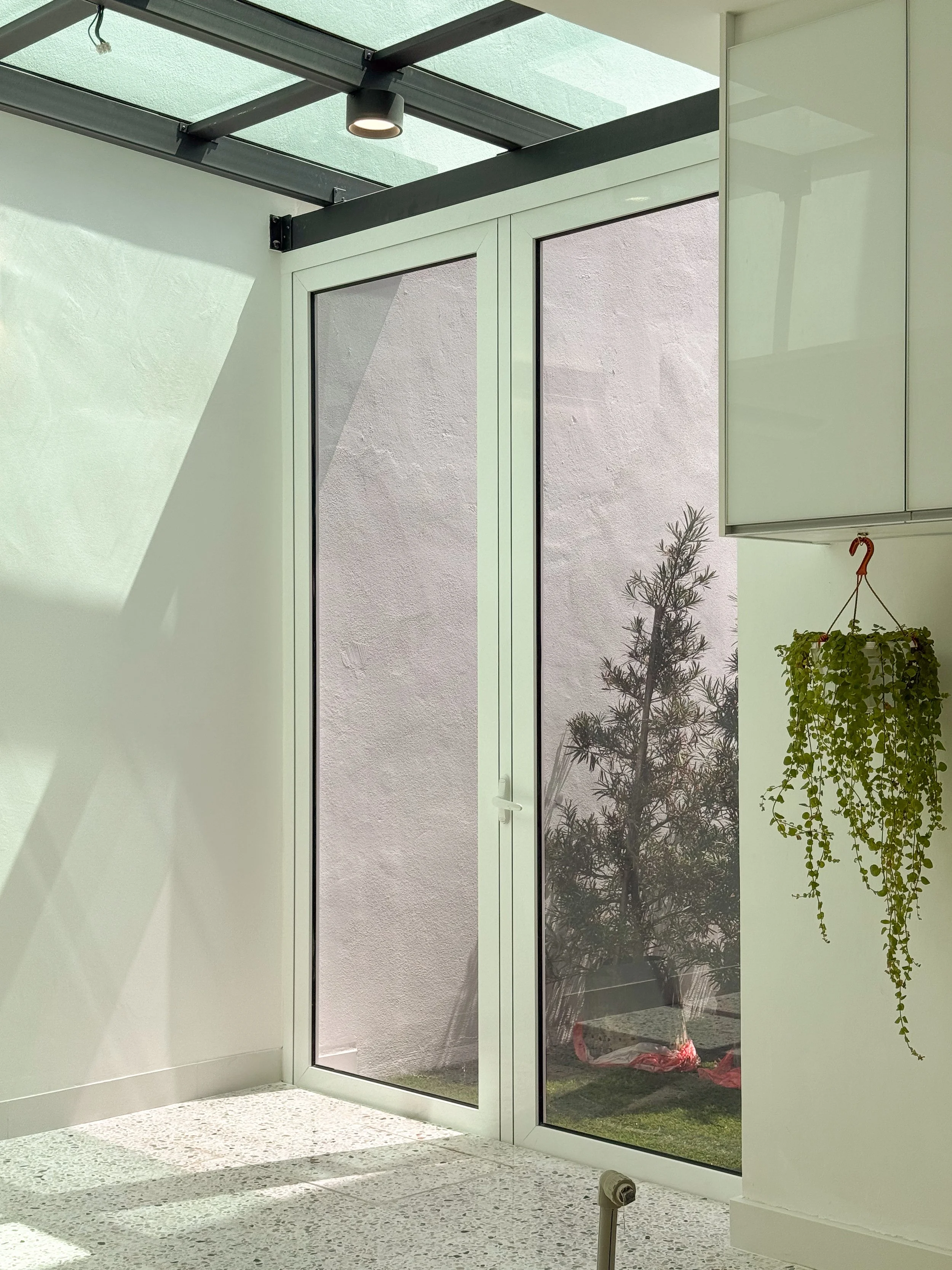 Interior of a modern room with glass doors, a glass ceiling, a potted plant hanging on the right, and sunlight casting shadows on the floor.
