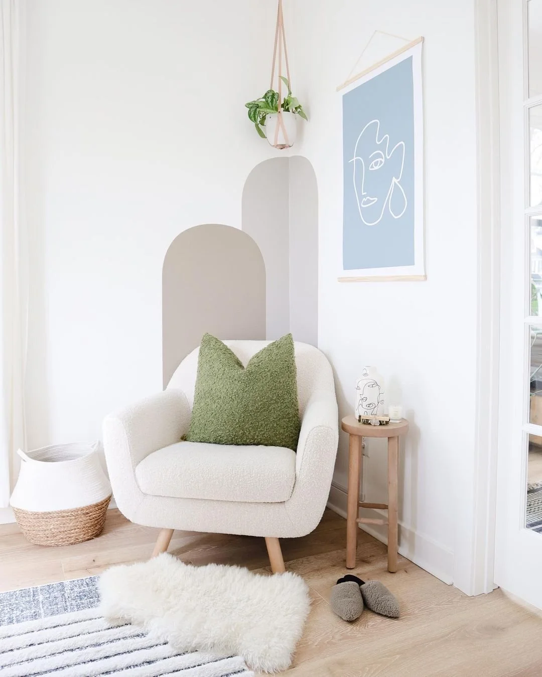 Cozy white armchair with green pillow in a modern living room corner, featuring decorative wall art, hanging plant, small wooden side table with candles, and woven basket on wooden floor.