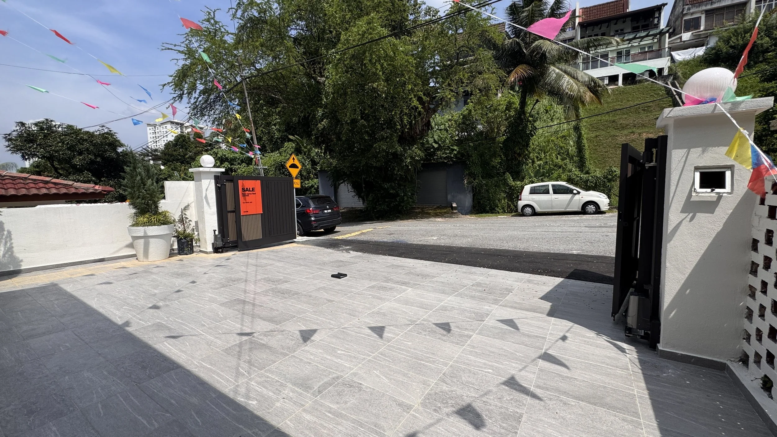 Driveway with a gated entrance, for sale sign on the left gate, small plants in pots, colorful triangular flags hanging overhead, and cars parked on the street in a residential area with trees and houses.