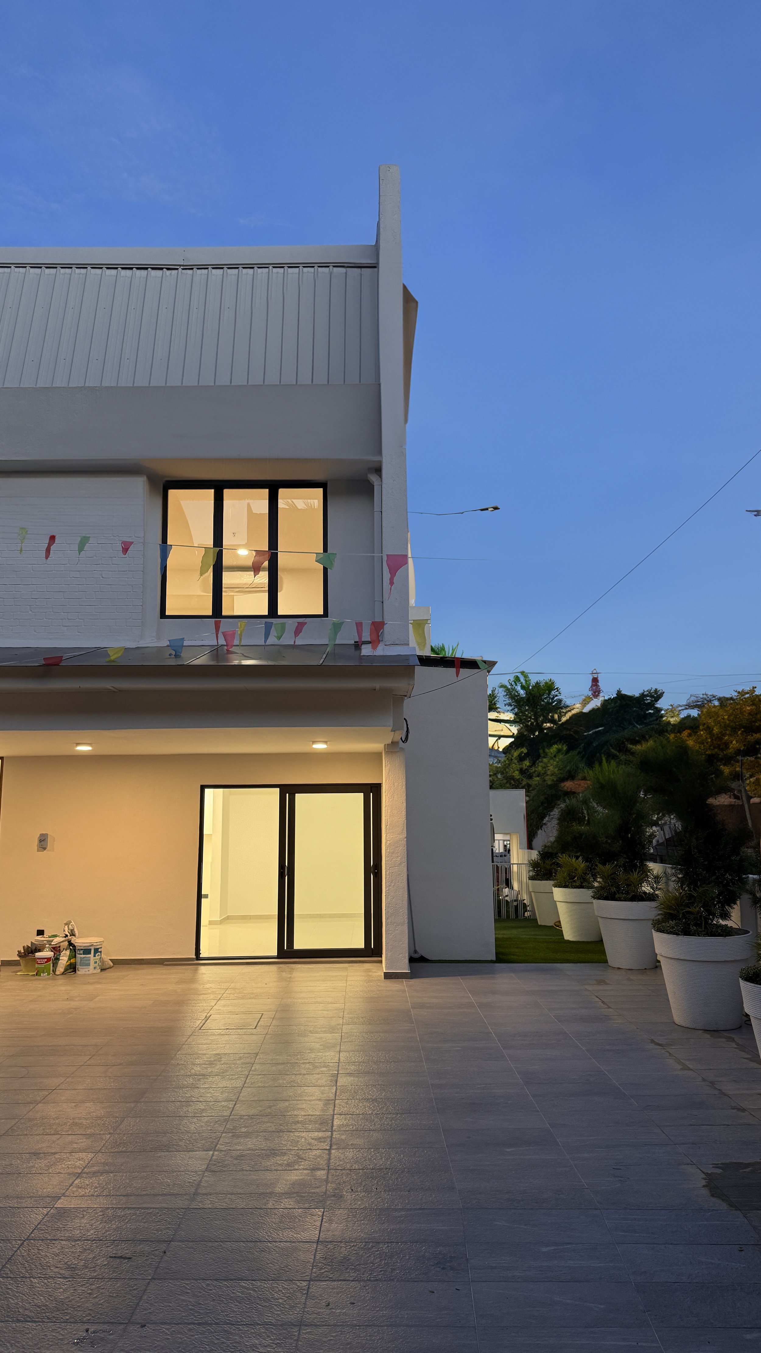 Modern house exterior with tiled patio, decorated with colorful flags and large plant pots, during twilight.