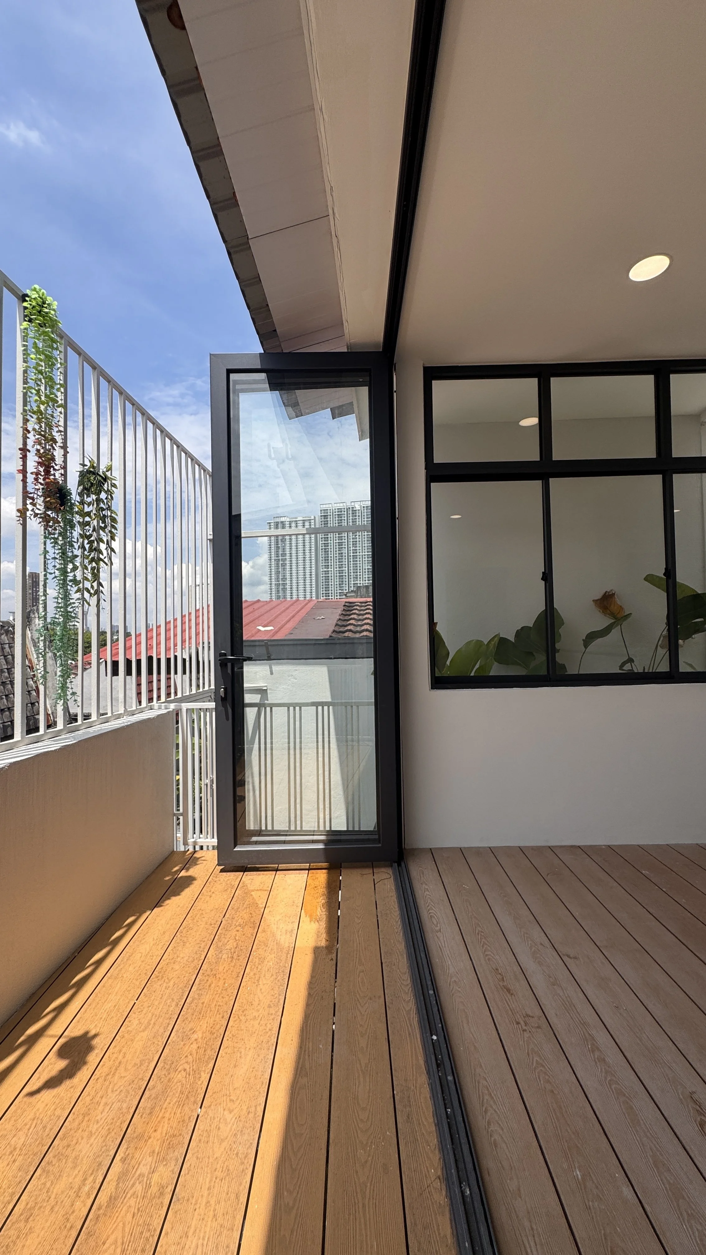 Modern balcony with wooden flooring, glass doors, black window frames, and outdoor plants.