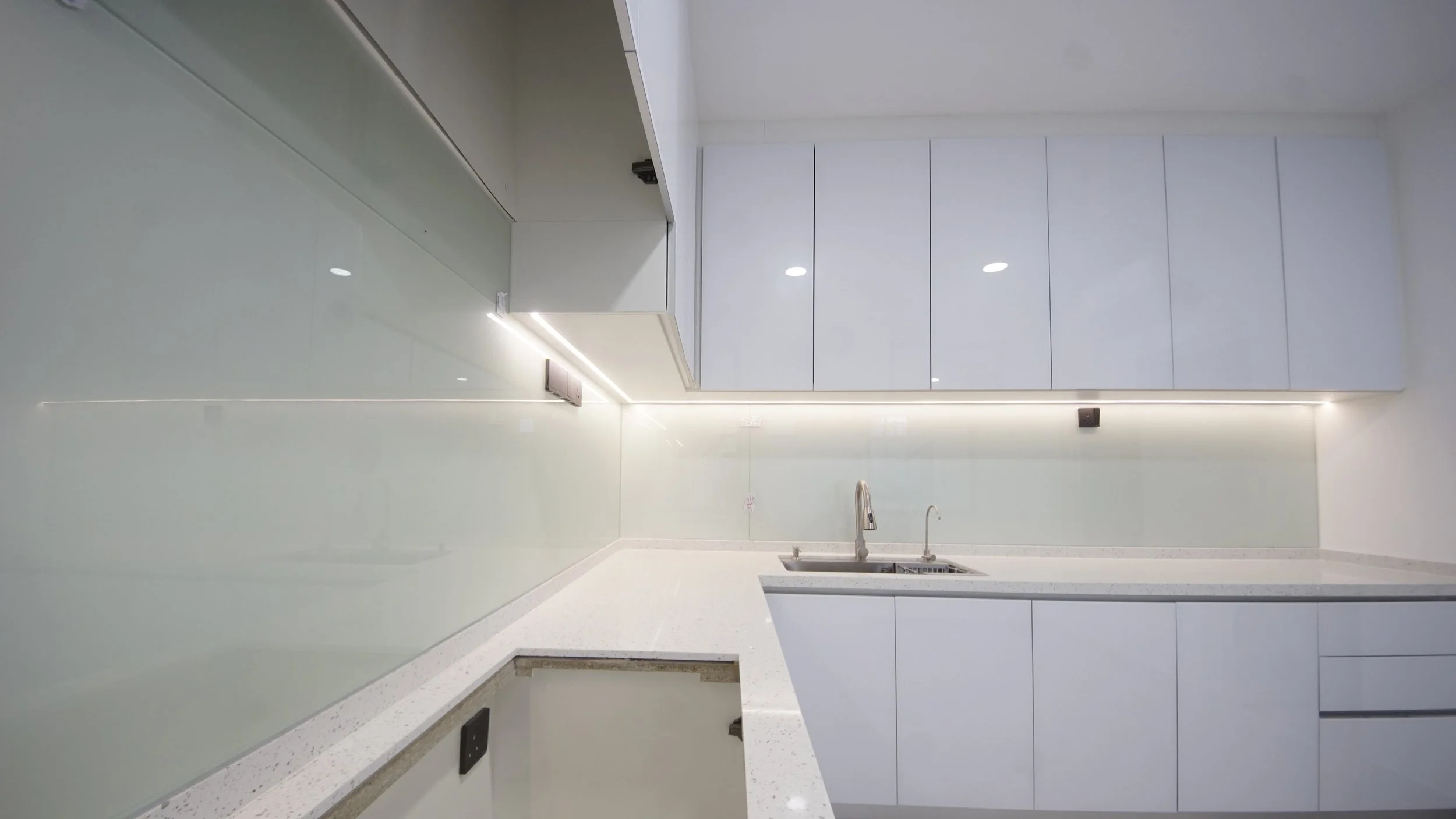 Modern kitchen with white cabinets, under-cabinet lighting, and a sink with a faucet on a white countertop.