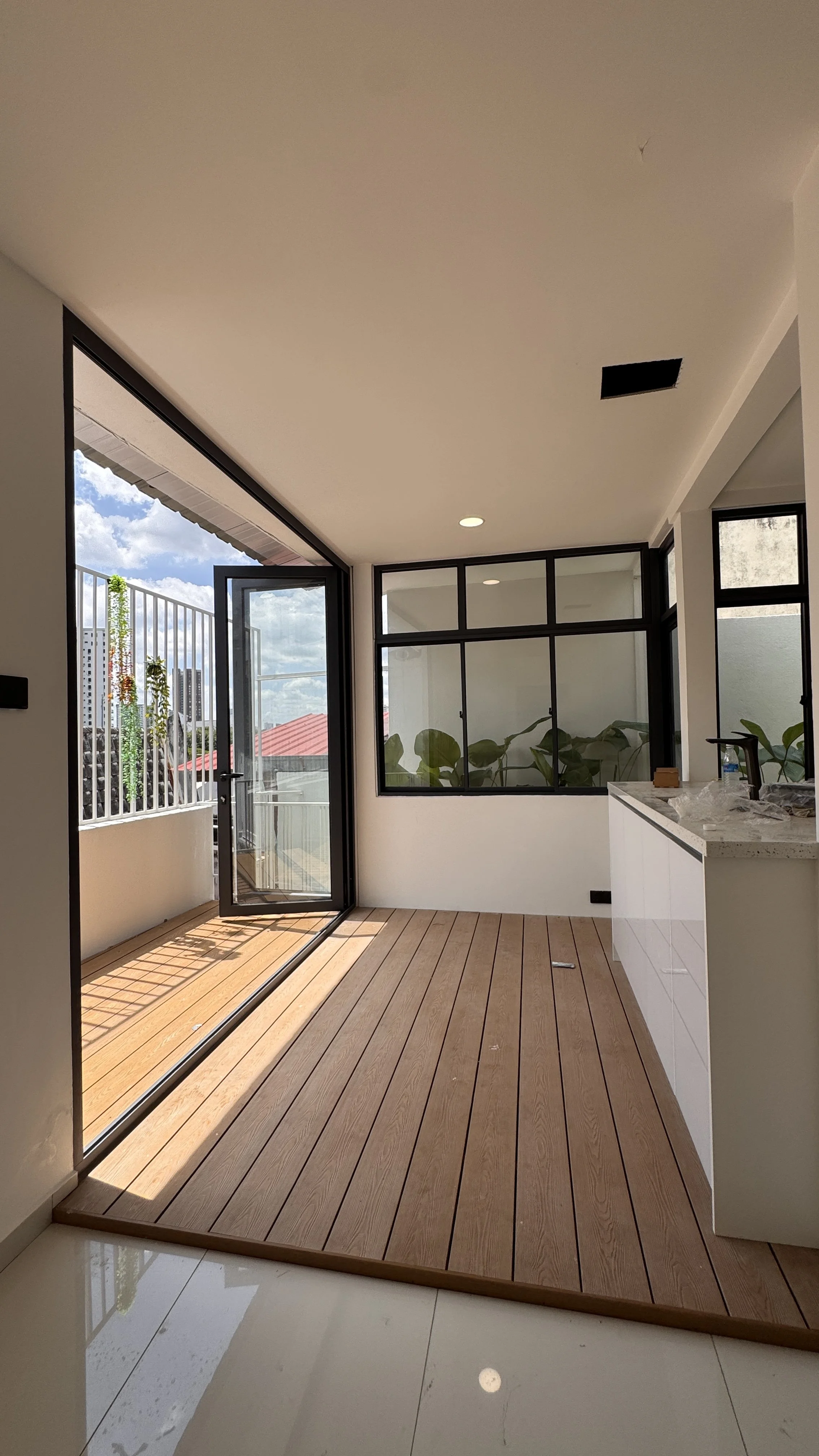Modern room with glass doors open to a balcony, wooden floor, indoor plants, and a countertop with a faucet, overlooking a cityscape.