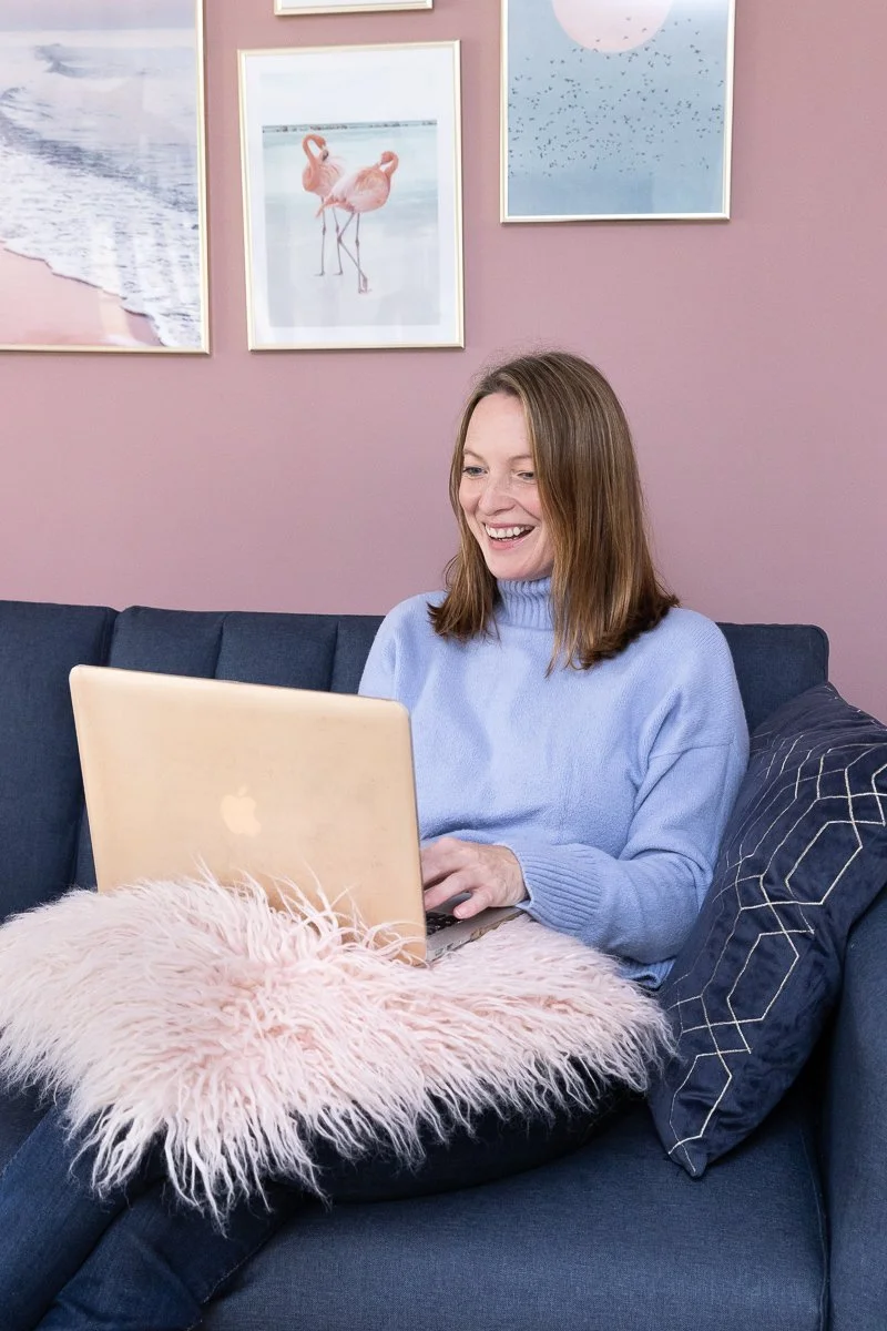 Woman sitting on a blue couch using a laptop, with a fuzzy pink cushion on her lap, and framed pictures on a pink wall behind her.