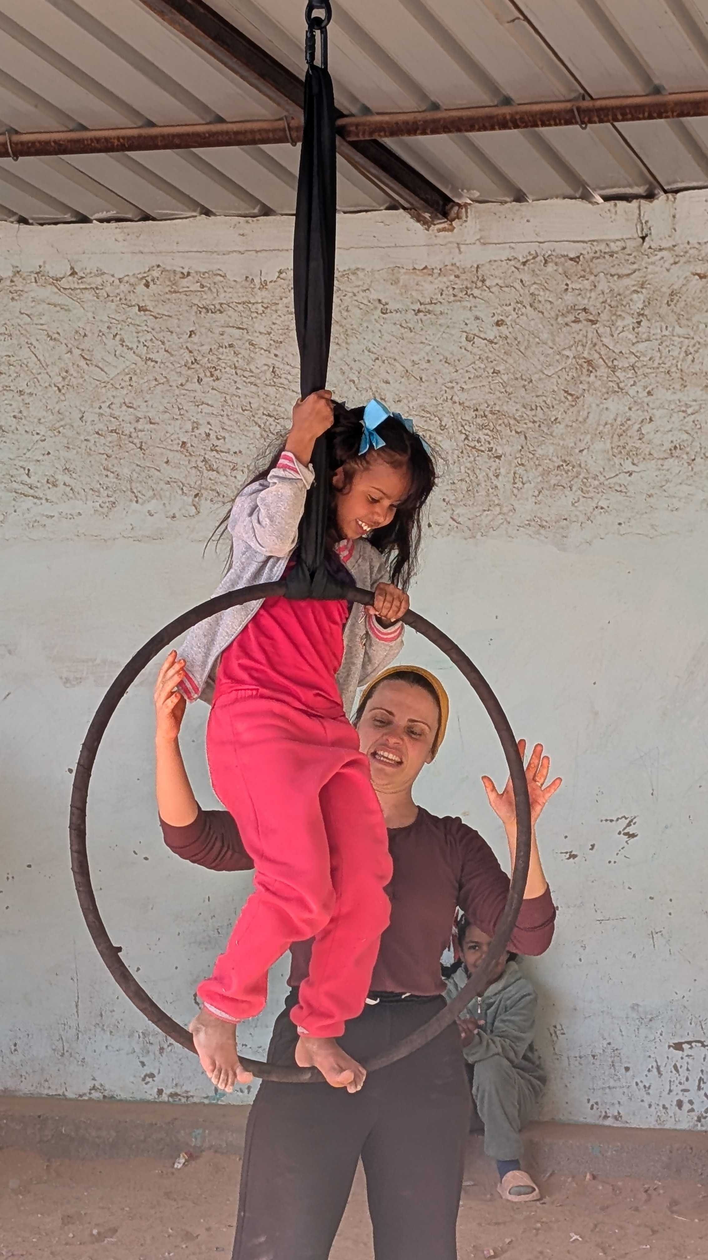 A woman is holding a young saharawi girl on a circular aerial hoop, with the girl smiling and holding onto the hoop. Another child sits on the ground in the background, looking on.