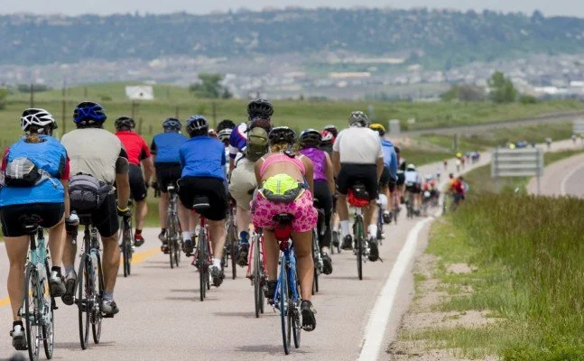 A group of cyclists riding on a rural road with green fields and hills in the background.