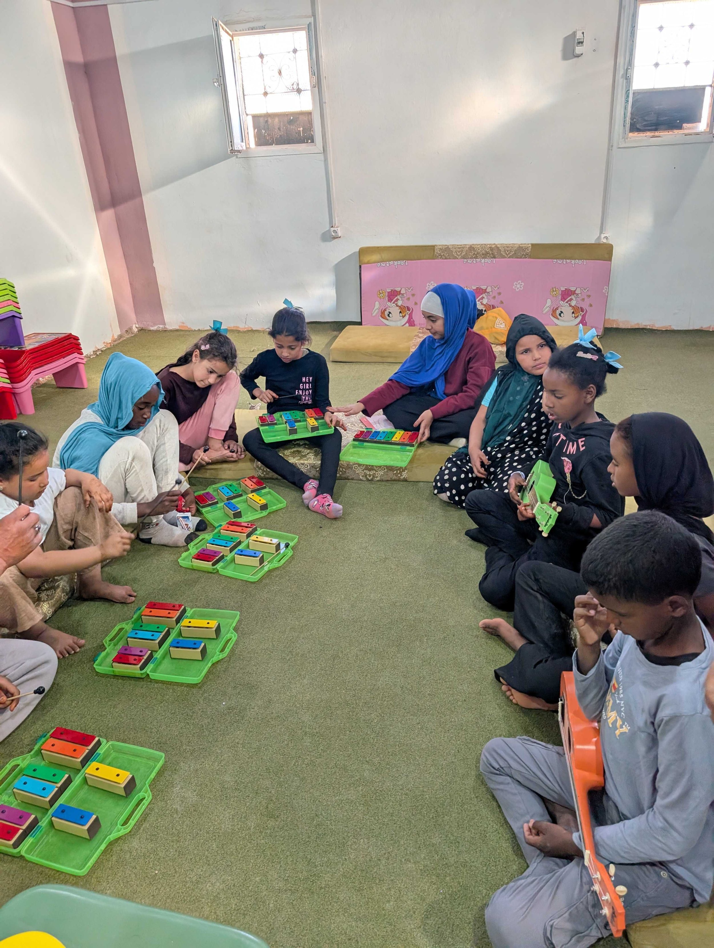 A group of saharawi children and an adult sitting on the floor, playing with colorful educational toys in a room with green carpet and white and pink walls, with small windows letting in natural light.