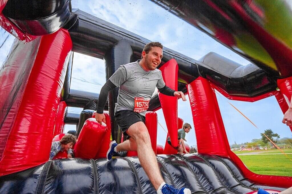 People participating in an obstacle course, climbing on a large inflatable structure resembling an obstacle with red and black sections, outdoors on a cloudy day.