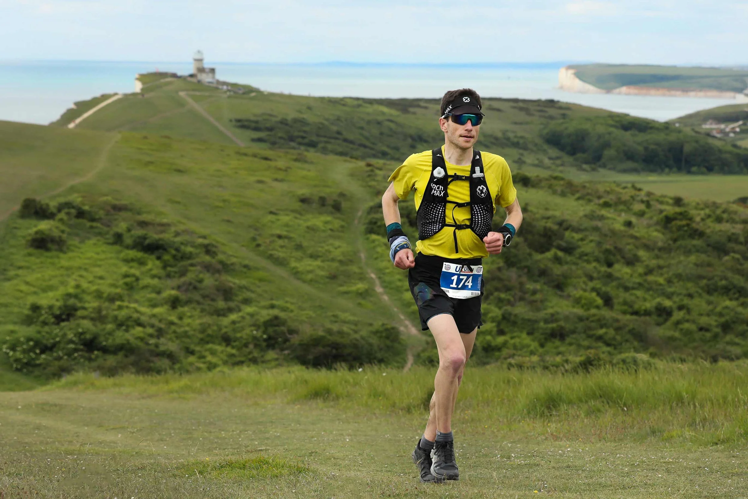 Male trail runner wearing a yellow shirt, black shorts, and a hydration vest running on a grassy trail with green hills and a lighthouse in the background.