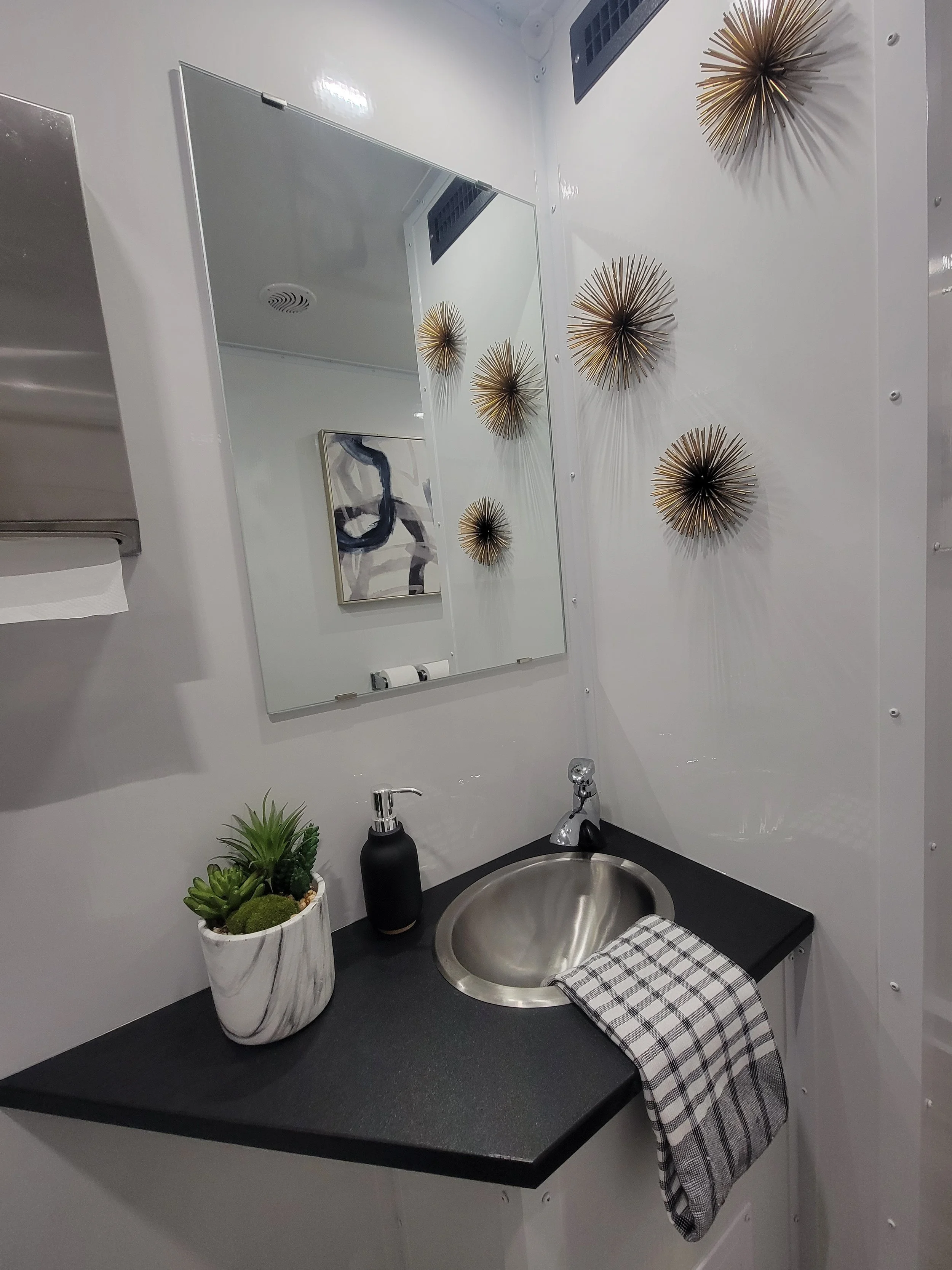 Modern bathroom with a countertop sink, decorative succulents in a marble pot, a black soap dispenser, and a checkered towel. The wall features gold starburst decorations and a large mirror.