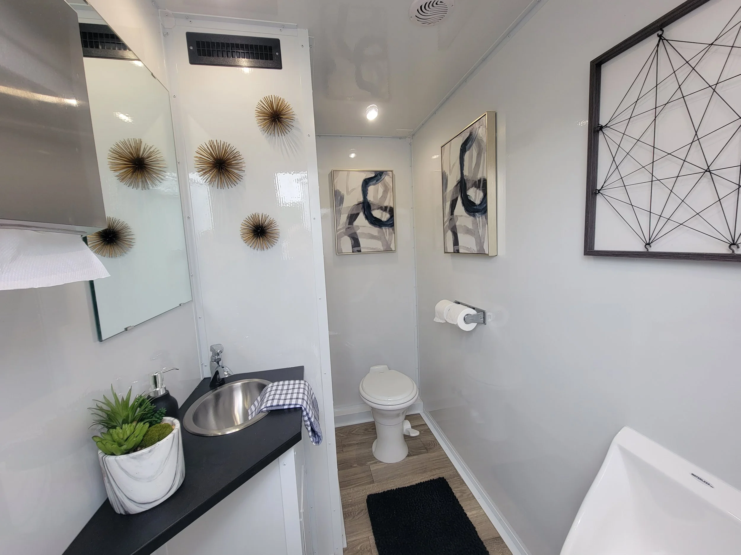 Modern restroom interior with sink, toilet, and urinal. Decor includes abstract art, geometric wall sculptures, and a potted succulent. Black countertop with checkered towel.