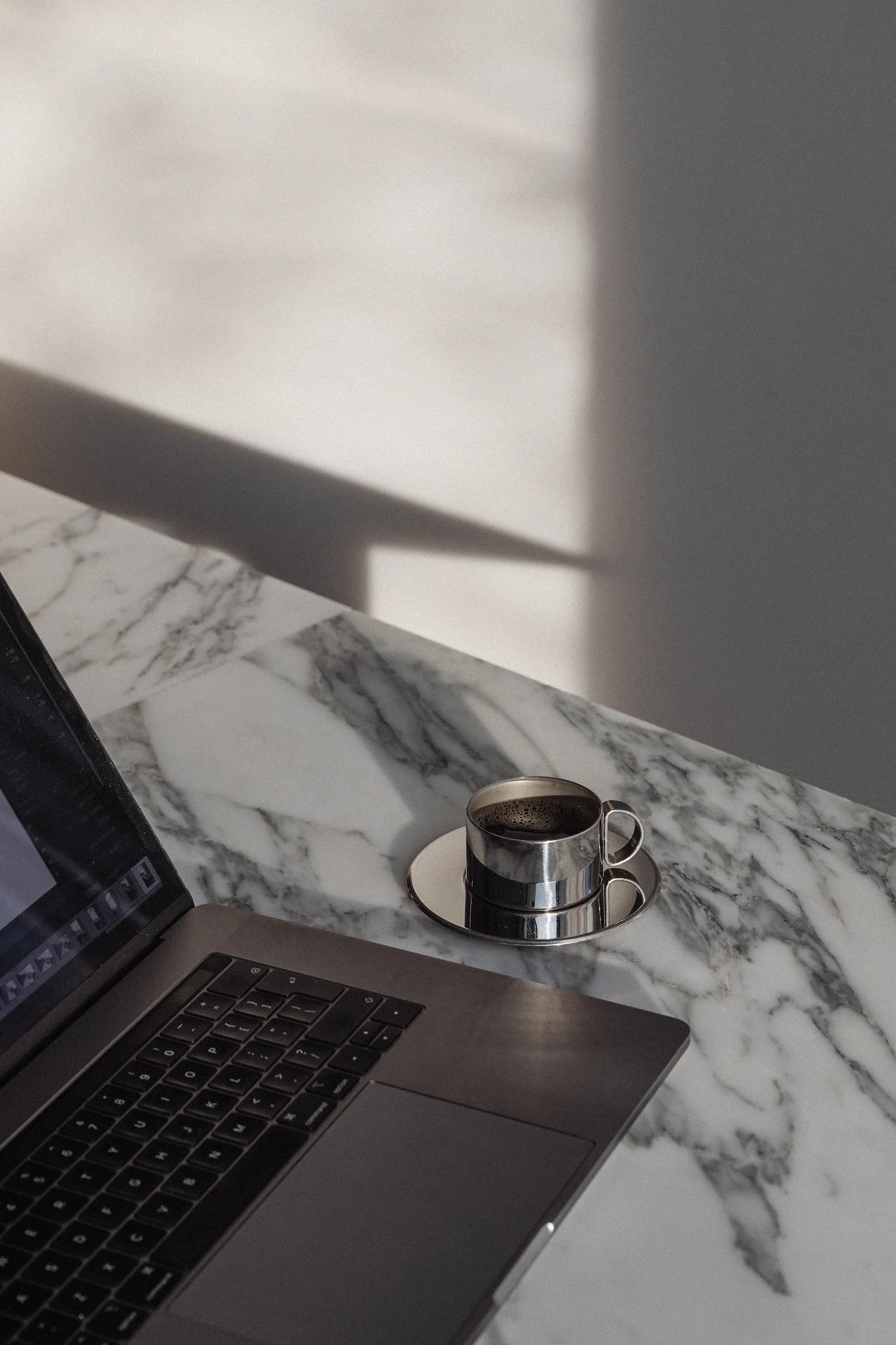 A marble countertop with a silver laptop and a black coffee in a silver cup on a matching saucer, with shadows cast on a white wall.