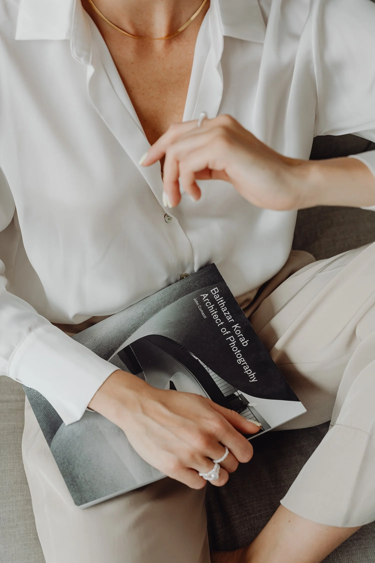 A woman dressed in white sits on a beige couch, holding a photography book titled 'Balhazar Korab Architect of Photography' by John Connolly. She is wearing rings and a delicate necklace.