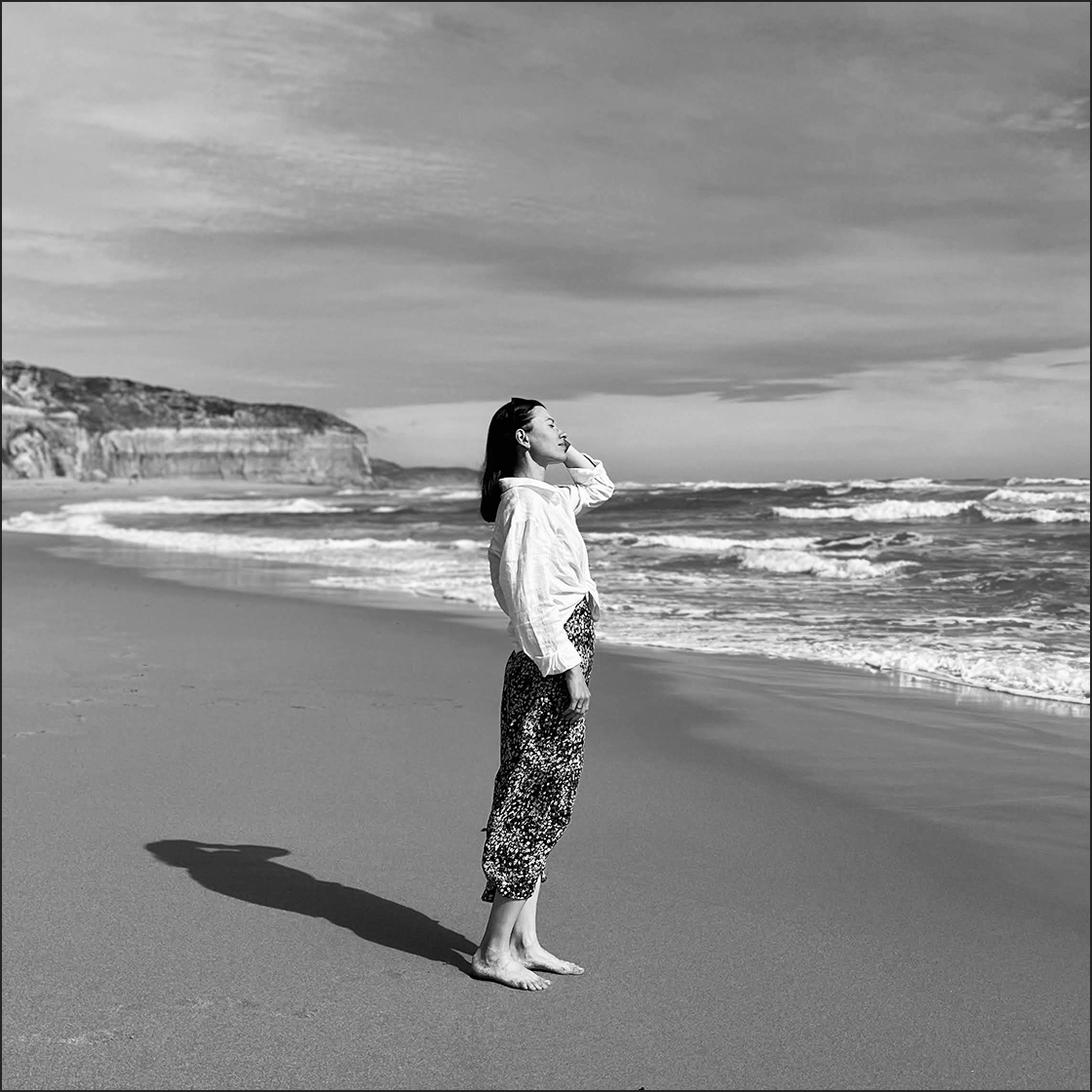 A woman standing barefoot on the beach, facing the ocean with her eyes closed, wearing a white shirt and patterned pants, with a shadow cast on the sand, under a cloudy sky.