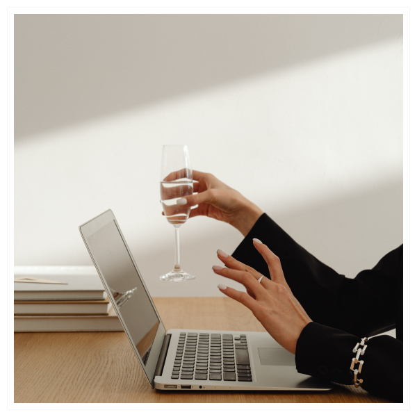 A person sitting at a desk, using a laptop, holding a glass of water in one hand, with a stack of books or notebooks on the desk.