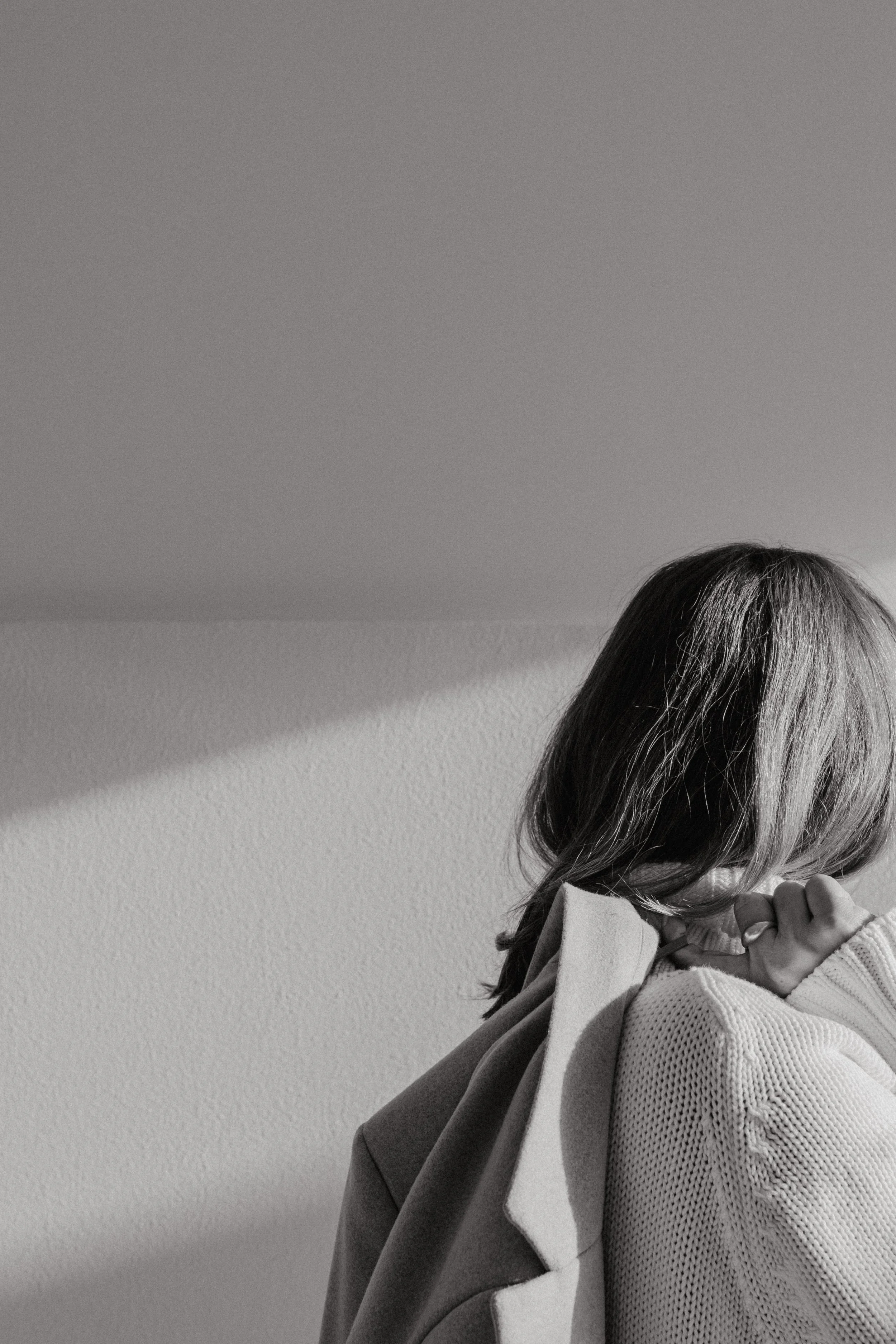 Black and white photo of a woman with shoulder-length hair, looking down with her hand touching her face, wearing a sweater and coat, against a plain wall background.