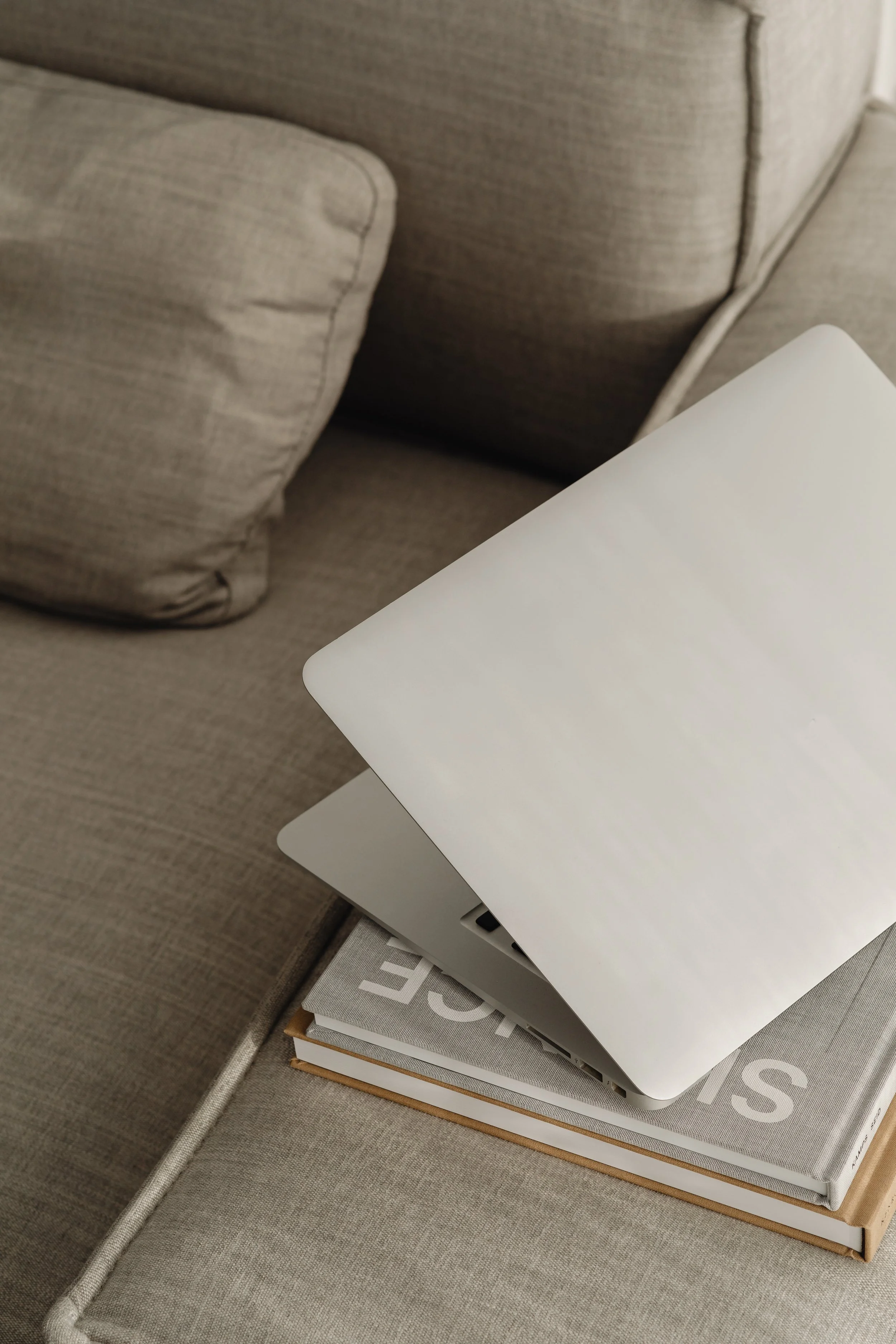 A closed silver laptop resting on top of two hardcover books with gray and white covers, placed on a beige fabric ottoman next to a beige couch.