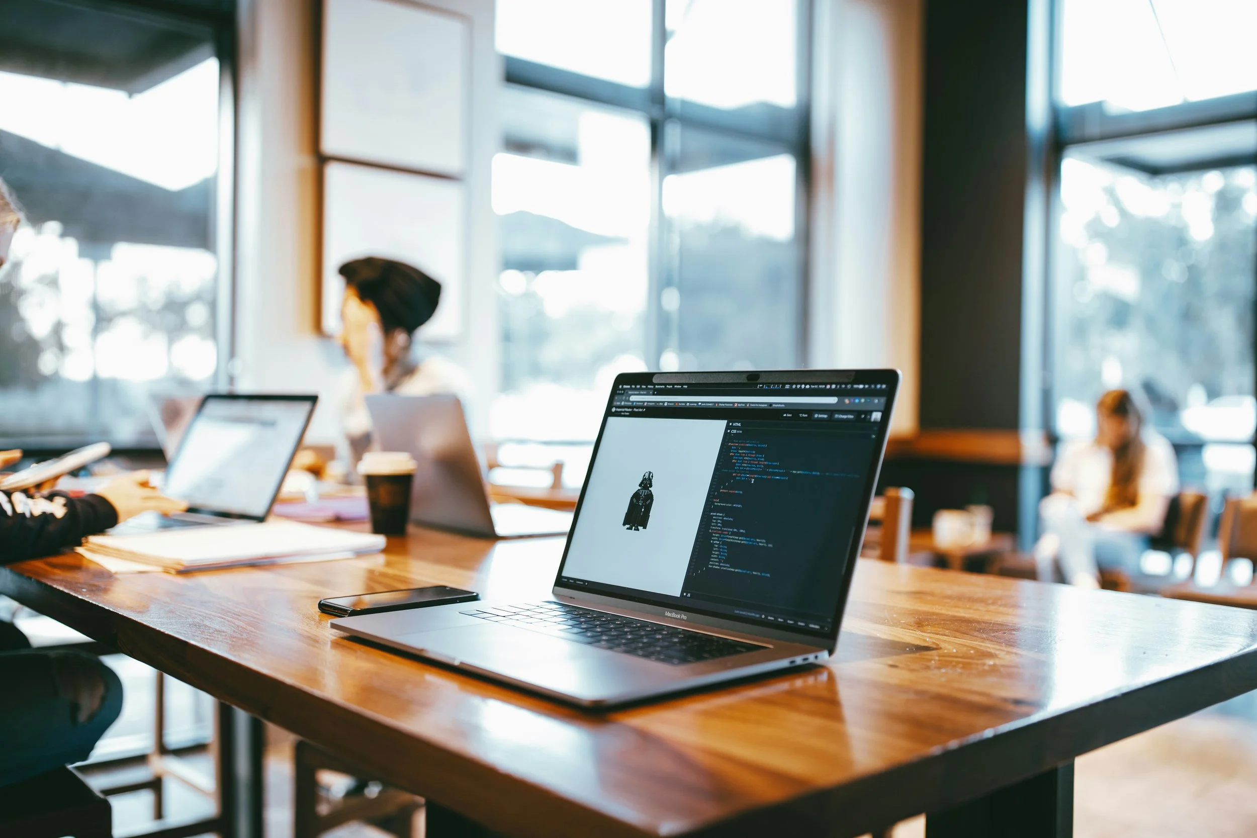 Laptop on a wooden table displaying a programming code and a small image of a person in a black garment. Two other laptops, a smartphone, papers, and coffee cups are on the table in a bright, modern cafe with large windows and people working.
