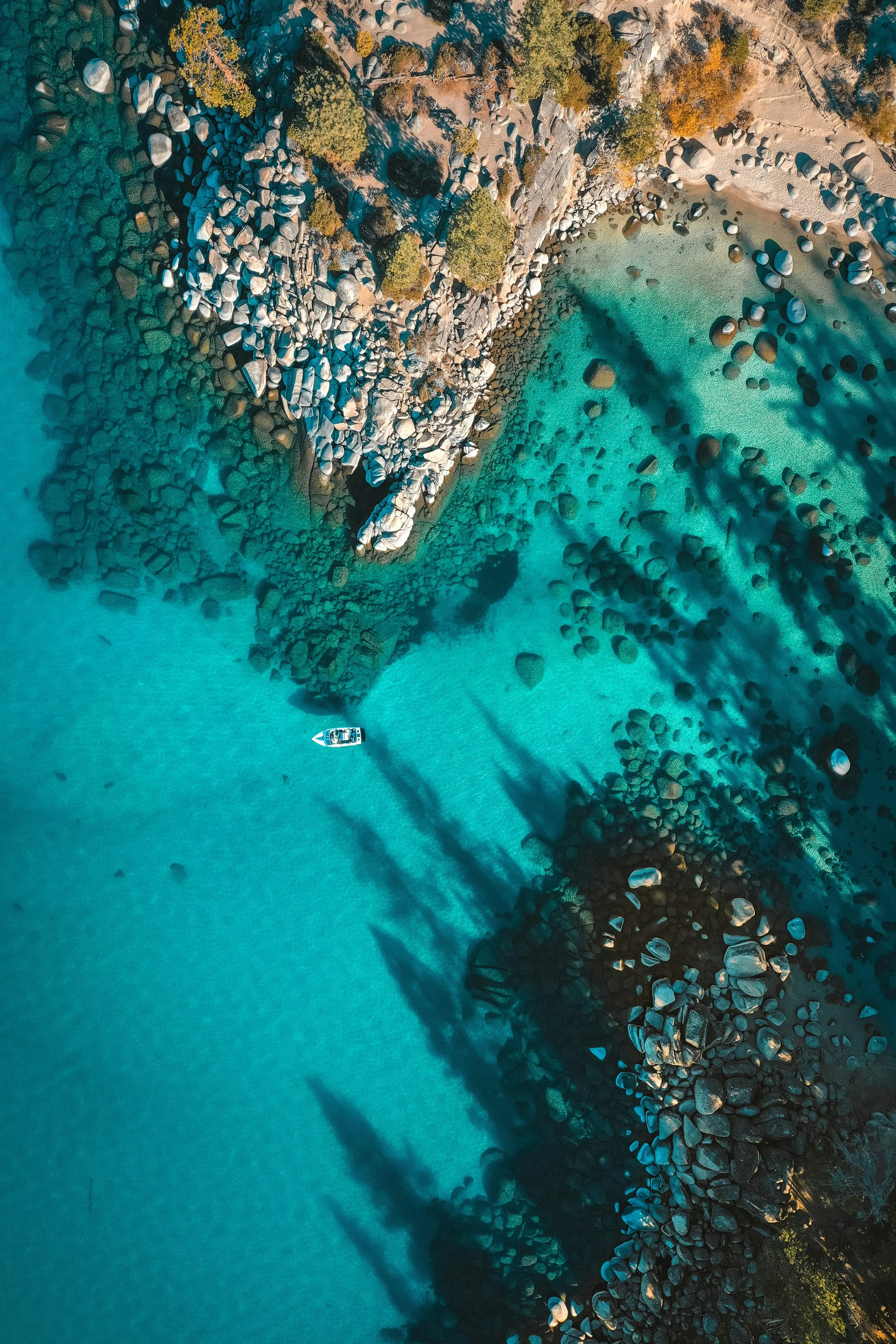 Aerial view of a boat in clear turquoise water near rocky cliffs with trees, casting shadows into the water.