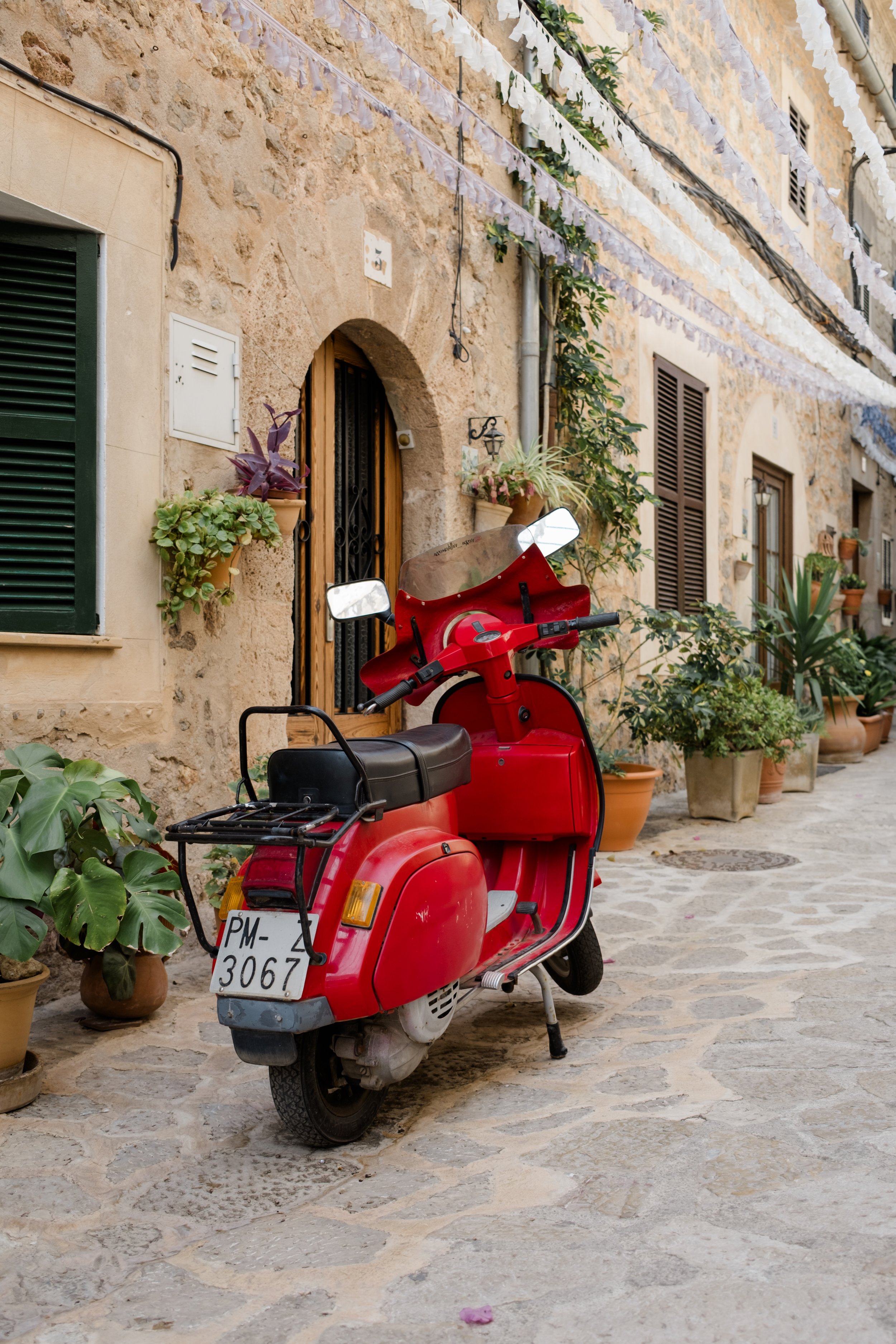 A red scooter parked on a cobblestone street in front of a stone building with potted plants and windows with shutters, under strings of white decorative flags.